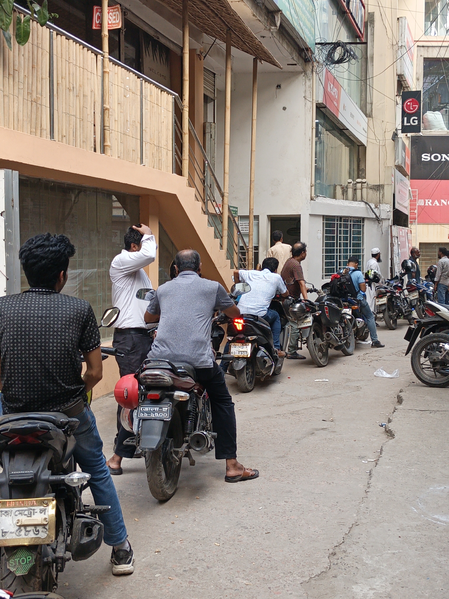 The image depicts a street scene in Dhaka, Bangladesh. A line of motorcycles is parked on a concrete street with various people near and on the motorcycles. The street is lined with buildings. Some buildings feature bamboo structures, signage, and exterior staircases. Visible signs include "CLOSED", "WELCOME", "LG", "SONY", and "RANGS". License plates with Bengali script are seen on the motorcycles. A cluster of tangled wires hangs above. The overall environment is urban, and the image provides a glimpse into daily life and transportation habits in Dhaka.