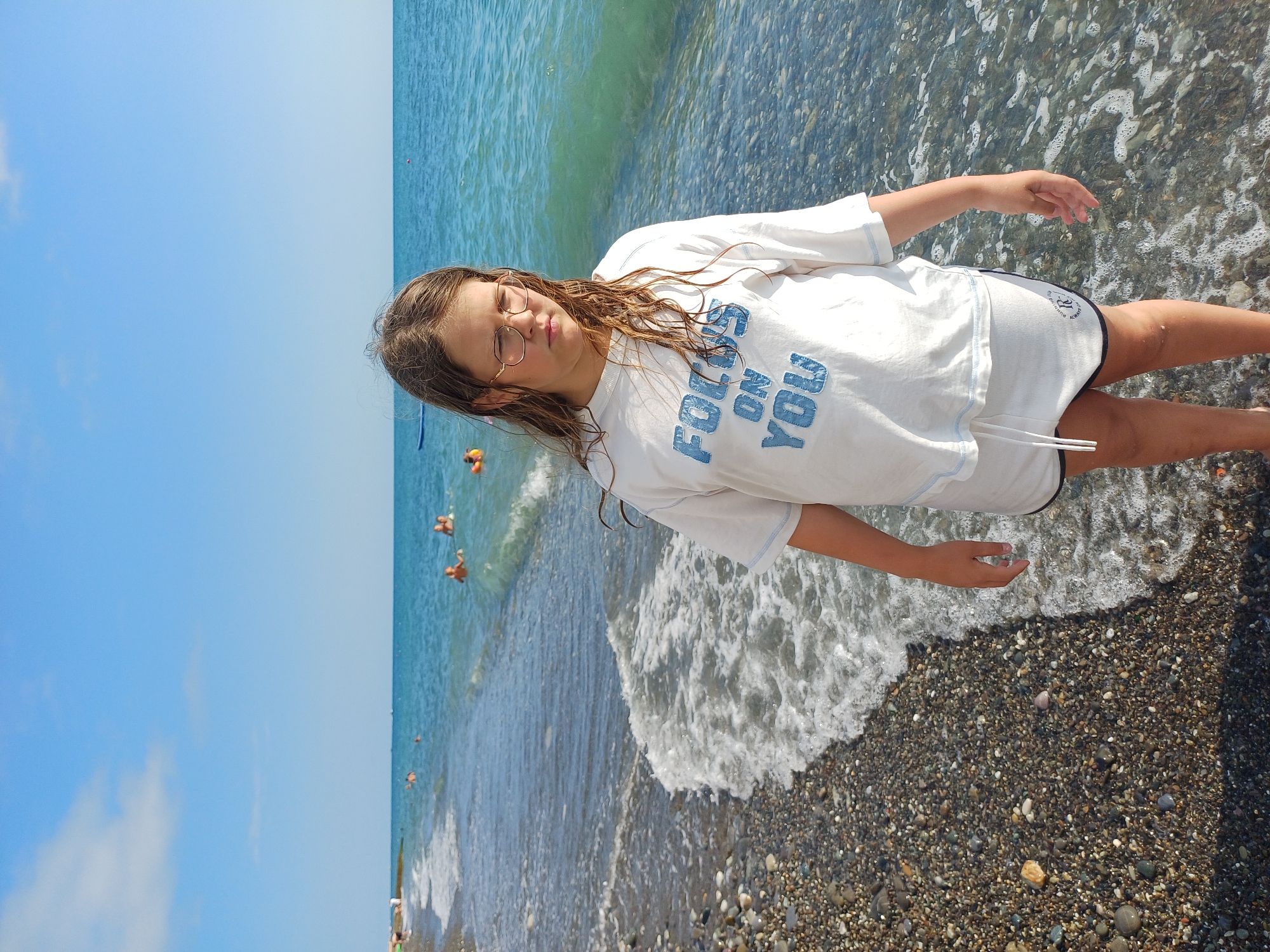 This image depicts a young girl standing at the edge of the sea on a pebble beach. She is wearing a white t-shirt with blue text that reads "FOCUS ON YOU" and white shorts. The girl has long, wet, brown hair and is wearing glasses. Her gaze is directed downwards, and she appears to be contemplating or in a relaxed state. The sea is a vibrant blue-green, with gentle waves washing ashore and creating foamy patterns on the pebbles. The sky above is a clear, bright blue with a few wispy clouds. Several other people are visible in the water in the distance, appearing to be swimming or enjoying the sea. The lighting suggests it is a sunny day, likely during daylight hours. The presence of the sea, pebbles, and the general atmosphere points to a beach location. The location context indicates this scene is in Sochi, Russia.