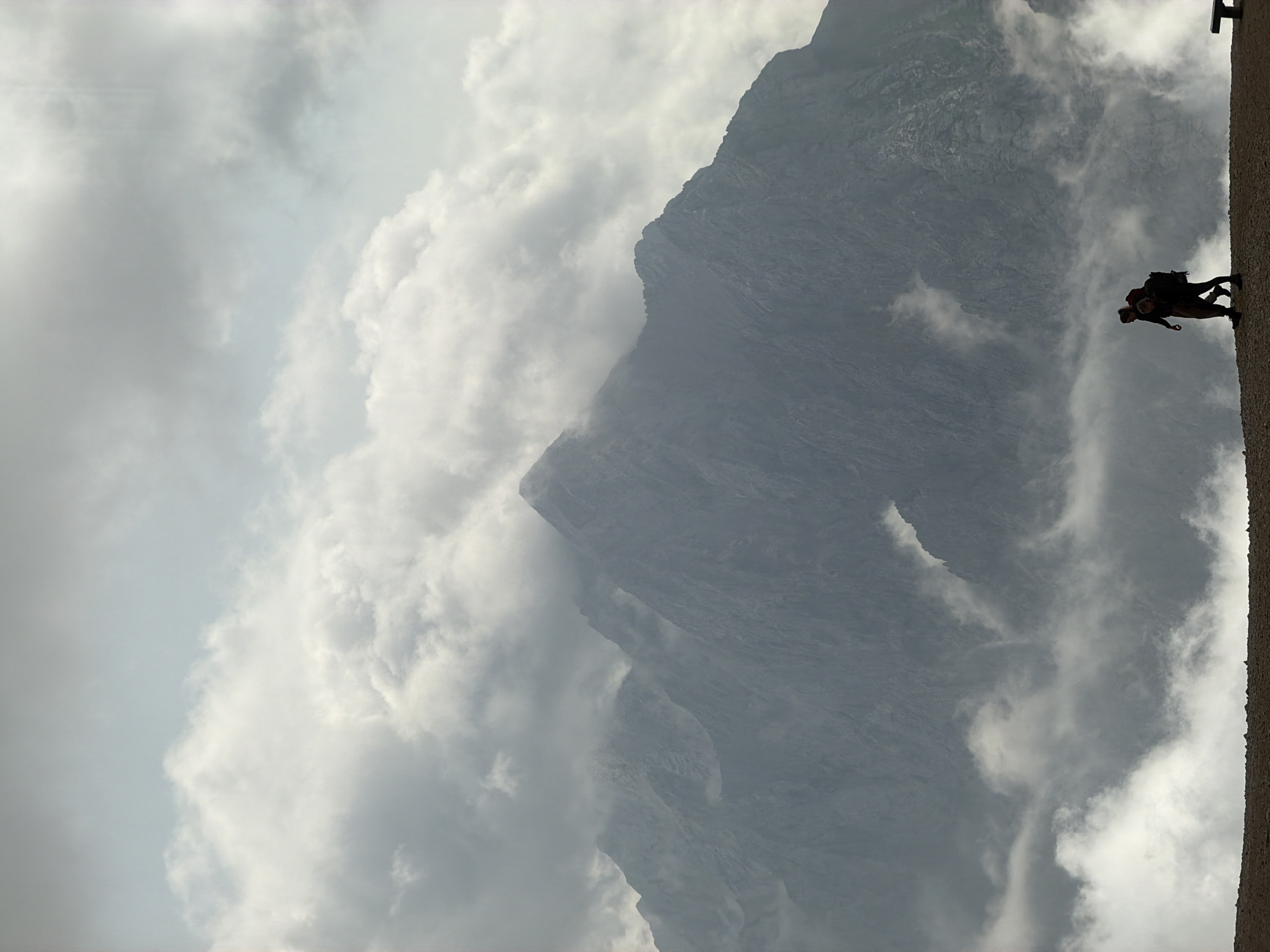 Two hikers are silhouetted against a dramatic mountain backdrop in Wildermieming, Austria. The hikers, one slightly behind the other, are walking from left to right along a gravelly path. Both are wearing backpacks, suggesting they are on a trek. The larger mountain range is shrouded in mist and clouds, with its peaks barely visible through the atmospheric conditions. The sky above is a mix of light and dark clouds, contributing to the moody and dramatic feel of the scene. The foreground is a rocky, earthy surface, while the background is dominated by the imposing, mist-covered mountains. A bench is visible on the far right of the image.