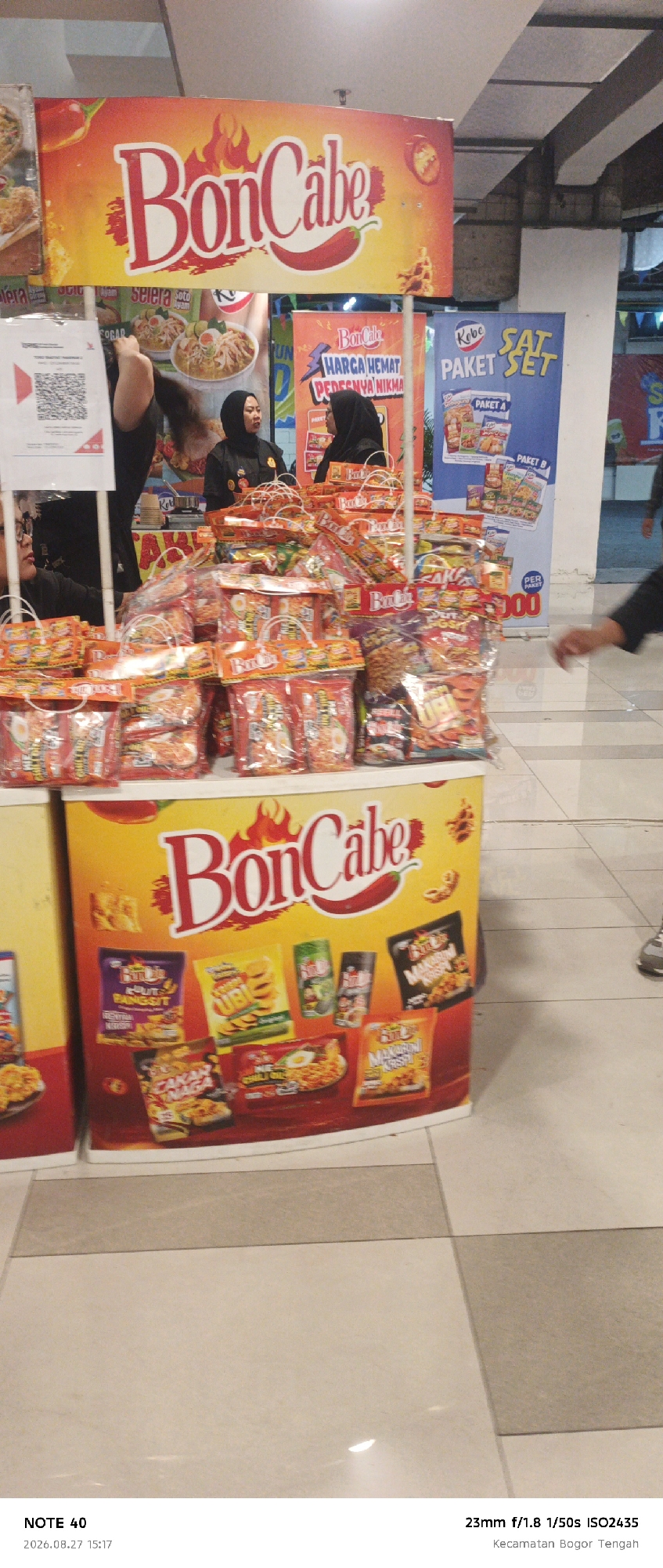 The image shows a stall selling BonCabe products, which appear to be spicy instant noodle seasonings and snacks. The stall is set up in what looks like an indoor shopping area, possibly a mall. There are multiple stacks of packaged BonCabe products on display. Behind the stall, two women wearing hijabs are visible, likely employees. One woman is standing and looking forward, while the other is partially obscured and appears to be interacting with a customer. A large banner with the BonCabe logo and product imagery is displayed prominently above the stall. Another banner to the right advertises "Paket Set" (Set Package). The floor appears to be tiled. The bottom of the image includes a watermark with "NOTE 40" and the date "2026.08.27 15:17", and camera settings "23mm f/1.8 1/50s ISO2435", along with the location "Kecamatan Bogor Tengah", indicating the scene is in Bogor Tengah District, Bogor, Indonesia.