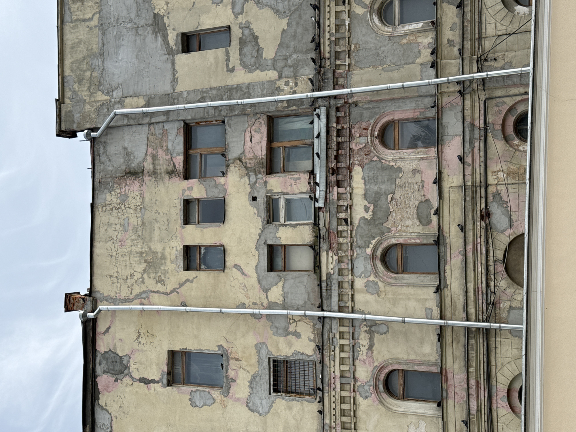 The image shows the exterior of an old, weathered building in Moscow, Russia. The building's facade is characterized by peeling paint, exposing brick and plaster underneath, and has a generally dilapidated appearance. Several windows are visible, some with barred coverings. Pigeons are perched on various ledges and windowsills of the building, and a few are seen in flight against a cloudy sky. Two downspouts run vertically along the facade, and a brick chimney is visible on the rooftop. The architecture suggests a historical building, possibly residential or an older commercial structure.