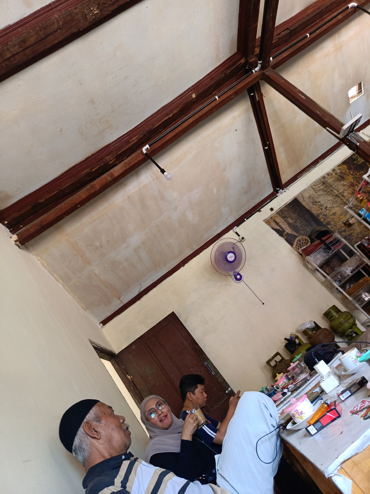 The image depicts an interior scene, likely a room in Kebonagung, Indonesia, characterized by exposed wooden beams supporting the ceiling. In the foreground, an elderly man wearing a black skullcap and a striped shirt is visible from the shoulders up, looking to the left. To his right, a woman wearing a light brown hijab and glasses smiles, facing slightly away from the camera. Beside her, a young man in a dark blue shirt is holding a glass of beverage and looking at his phone. They are seated at a table laden with various items, including cups, plates, lighters, and what appear to be cigarette packs. In the background, a doorway leads to another area, and a purple oscillating fan is mounted on the wall. Further into the room, a patterned rug covers the floor, and a shelf unit holds several items, including gas canisters and other household objects. The overall atmosphere suggests a casual gathering or a moment of rest.