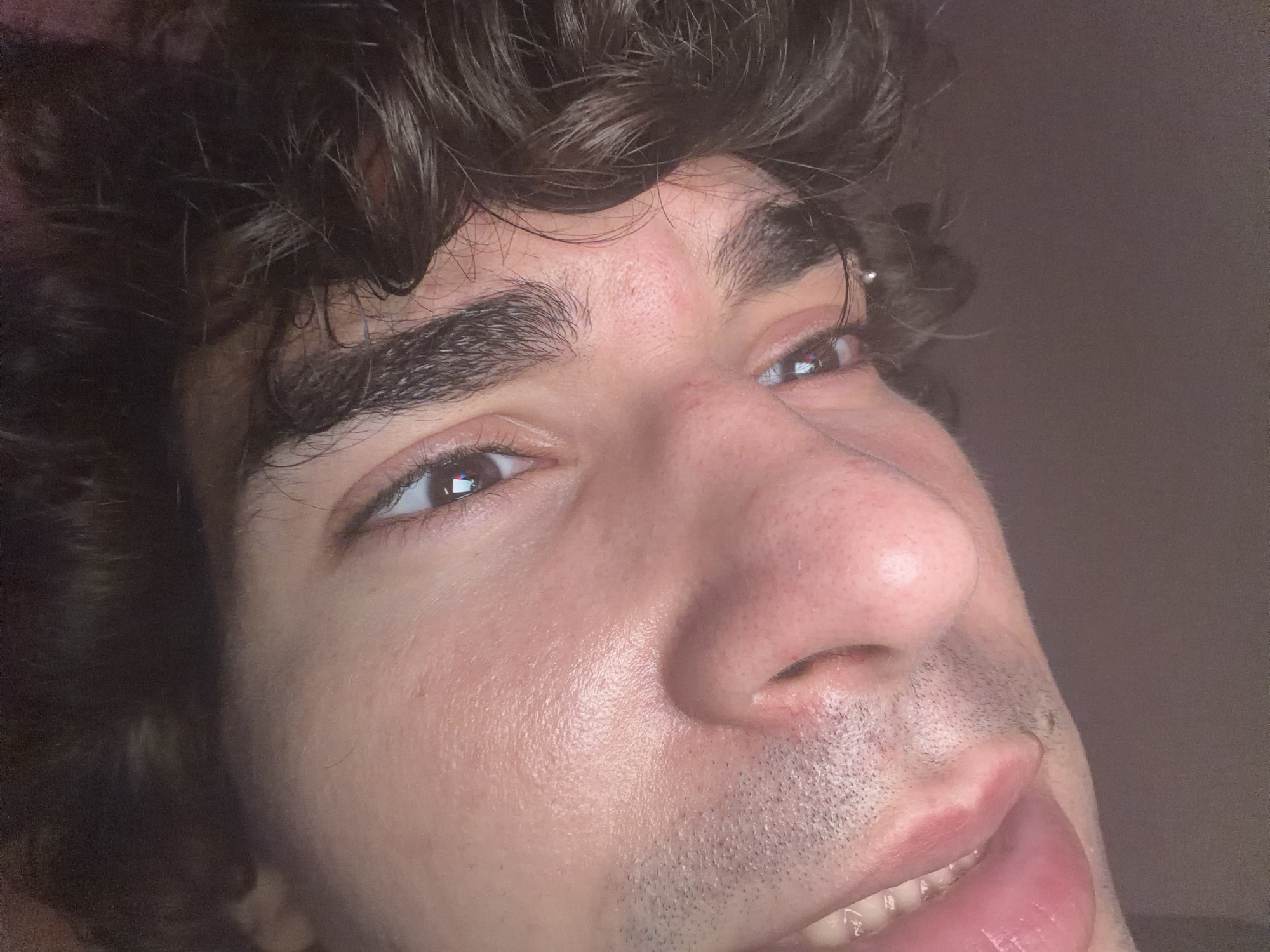 This close-up, angled photograph captures the face of an individual. The subject has dark, curly hair visible at the top and left of the frame, falling across the forehead. Thick, dark eyebrows frame brown eyes, each reflecting distinct light sources, possibly from a screen or window. A small, shiny piercing is visible near the outer corner of the right eye (viewer's right ear). The skin is light to medium in tone, exhibiting visible pores and a slight sheen across the forehead, nose, and cheeks, along with minor reddish marks on the forehead and bridge of the nose. A prominent nose is centered in the mid-frame. Short, dark stubble covers the upper lip, chin, and jawline. The mouth is partially open, revealing white upper teeth and the tip of a pink tongue. The lips are full and appear glossy. The subject's gaze is directed upwards and to the viewer's right. The background is a plain, uniform brownish-grey surface, offering no specific environmental details. The image provides no visual information to determine its specific geographical location, such as Sofia, Bulgaria.