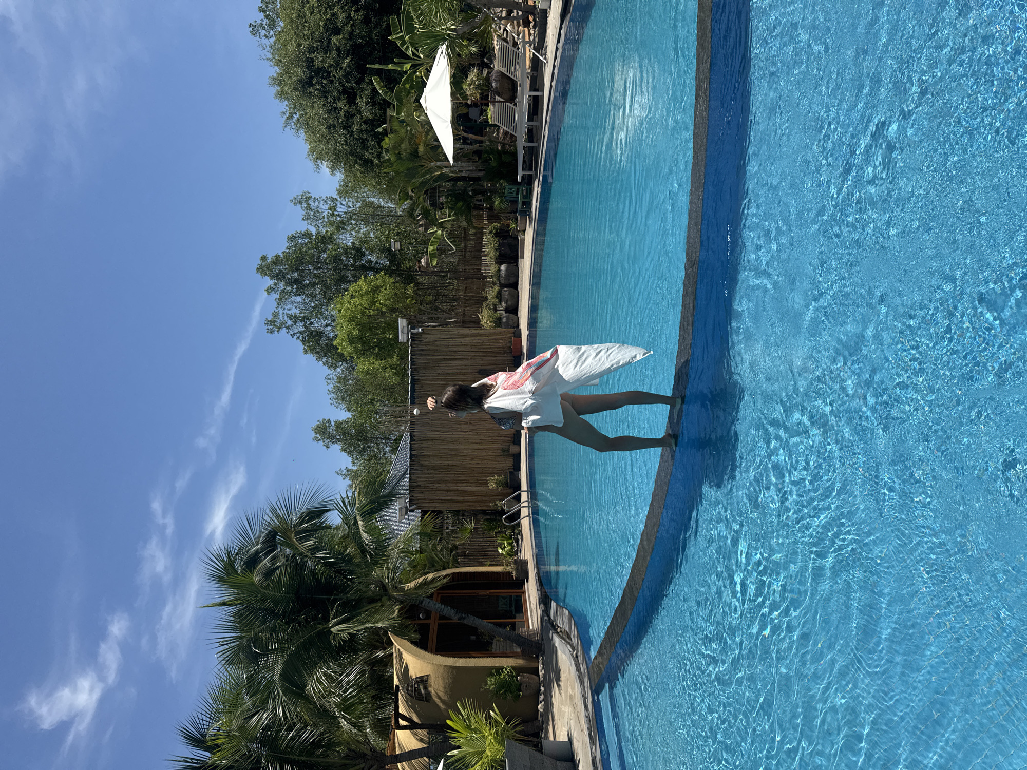 In Mũi Né, Vietnam, a woman stands on the edge of a vibrant blue swimming pool, her back to the camera. She is wearing a swimsuit and a light, flowing cover-up with a flamingo pattern. Her hair is pulled up, and she appears to be reaching towards her head with one hand. The pool is surrounded by lush greenery, including palm trees and other tropical plants. In the background, there is a uniquely shaped hut with a thatched roof, a bamboo fence, and lounge chairs with a white umbrella. The sky above is a clear, bright blue with a few wispy clouds. The scene captures a serene and idyllic vacation setting.
