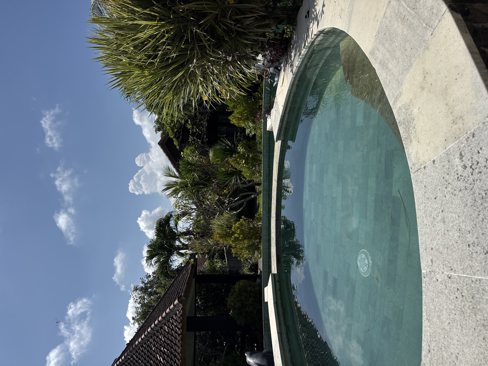 The image depicts a serene outdoor scene in Kuta Selatan, Indonesia. A circular swimming pool, lined with light-colored stone, dominates the foreground. The pool water reflects the sky, clouds, and surrounding vegetation. Tropical plants, including palm trees and lush foliage, surround the pool area. In the background, a traditional Balinese-style building with a terracotta-tiled roof is visible amidst the greenery. The sky is blue with scattered white clouds.