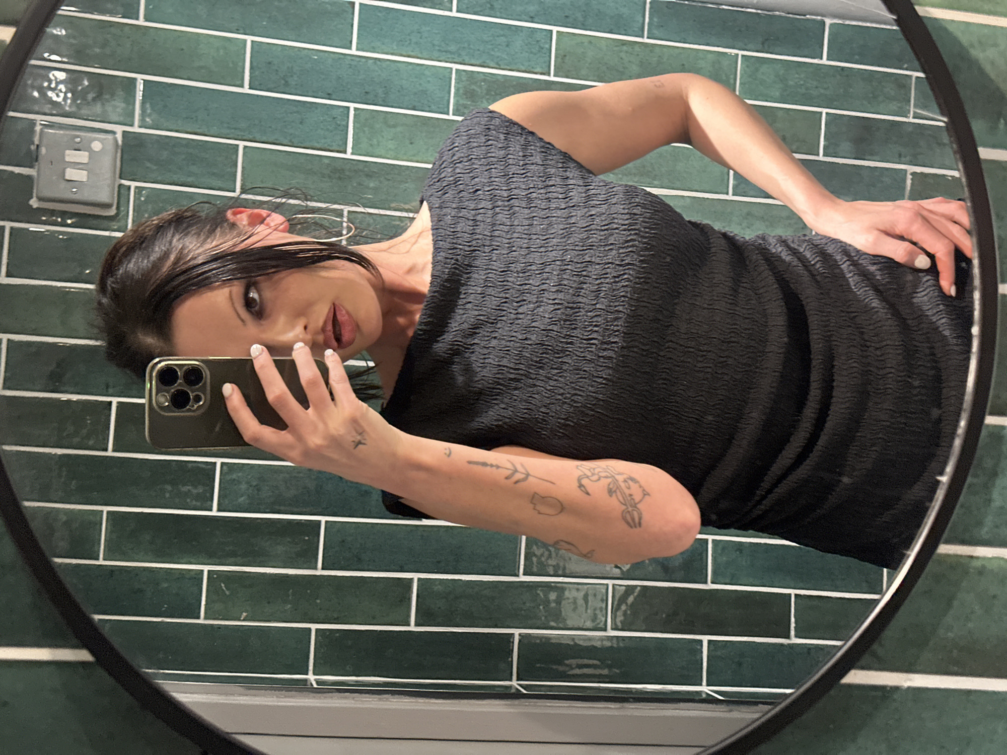 A female subject is depicted taking a mirror selfie indoors. She has dark hair, light skin, and wears a dark, textured smocked dress with a boat neckline. She accessorizes with gold hoop earrings and white nail polish. Her left arm features several small, black line-art tattoos, including a scorpion-like design and botanical elements, with another small tattoo on her left hand. The subject holds a dark-colored smartphone, an apparent iPhone Pro model, in her left hand, while her right hand rests on her hip. She looks towards her reflection with a slightly open mouth. The background, reflected in a round, black-framed mirror, consists of a wall tiled with vertically oriented, rectangular dark teal glazed tiles set with white grout. A grey double light switch, indicative of UK electrical fittings, is mounted on the tiled wall. The precise location within London, United Kingdom, cannot be ascertained from the image.