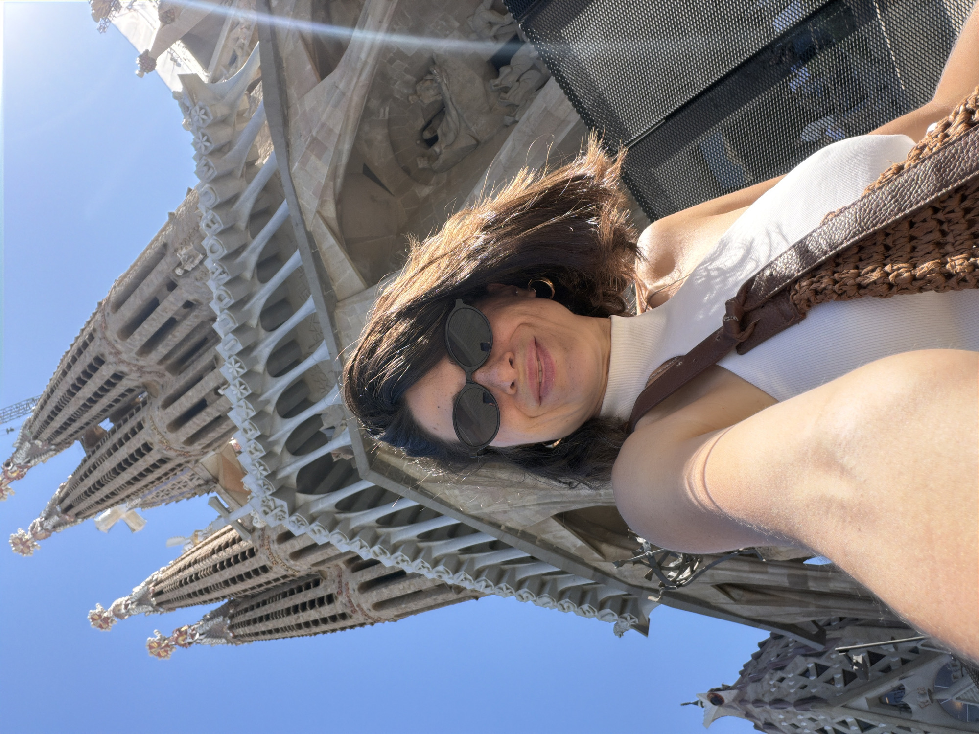 The image shows a woman taking a selfie in front of the Sagrada Familia in Barcelona, Spain. She is wearing sunglasses, a white sleeveless top, and a brown woven bag slung over her shoulder. The Sagrada Familia's distinctive spires and intricate facades are visible in the background, set against a clear blue sky. The sun is shining, casting a bright light on the scene. The perspective is from a low angle, looking up at the basilica.
