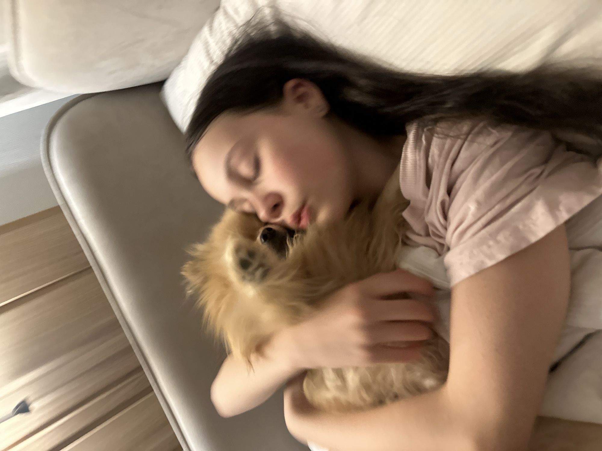 A young female with dark hair is lying in bed, embracing a Pomeranian dog. The girl's eyes are closed, and she appears to be sleeping or resting her head on the dog. The dog is light brown, with a fluffy coat. They are both on a bed with a white pillow. The location of Votkinsk, Russia, is not evident in the image.