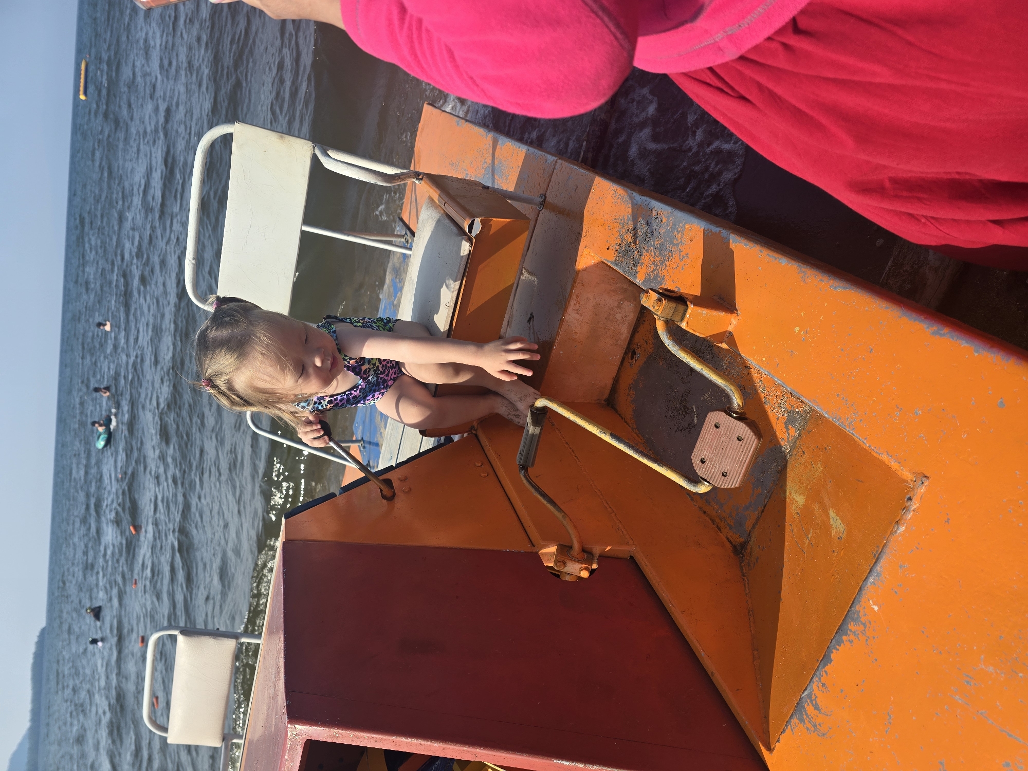 A young child with blonde pigtails sits on an orange pedal boat on a body of water. The child is wearing a colorful swimsuit and appears to be reaching down towards the boat's pedals. The boat has white seats and is partially submerged in the water. In the background, other people are visible in the water, some swimming and others on floatation devices. The sky is clear and blue, and there is a distant shoreline. The scene appears to be in Maksimiha, Russia, based on the context provided.