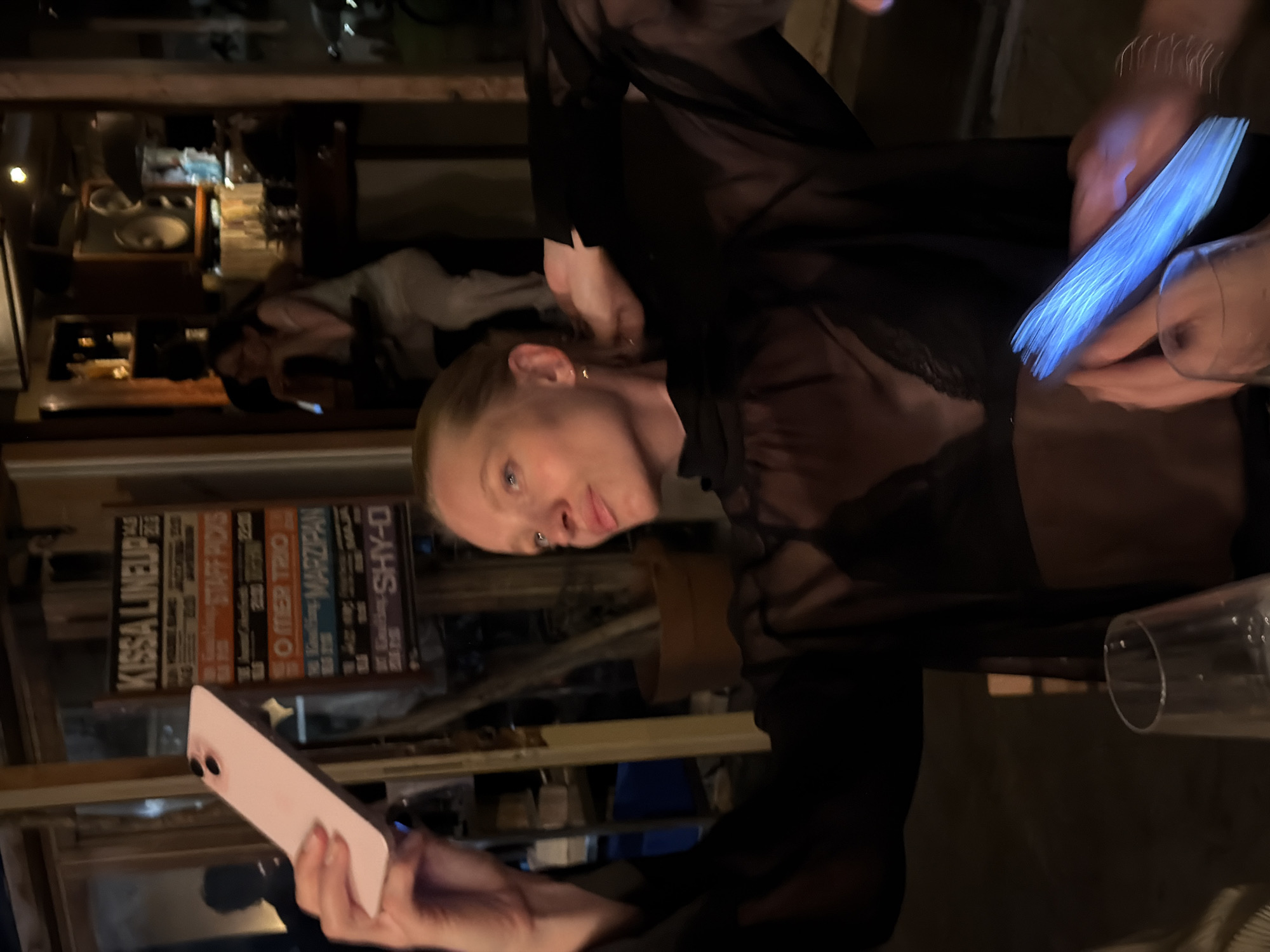 A blonde woman is taking a selfie with a pink phone in a dimly lit, indoor establishment. She is wearing a sheer black blouse and a black bra. The background shows a sign with text including "KISSA LINEUP," and other people in the background. The establishment appears to be a bar or restaurant. The sign is hanging on a wooden wall, suggesting a rustic or casual atmosphere. The lighting is warm and ambient, with some areas darker than others, indicating evening or nighttime. The woman's pose suggests she is aware of the camera and is engaging with it. The presence of the sign and the overall ambiance suggest this might be a venue in Tel-Aviv, Israel, possibly a music venue or bar, given the "LINEUP" text.