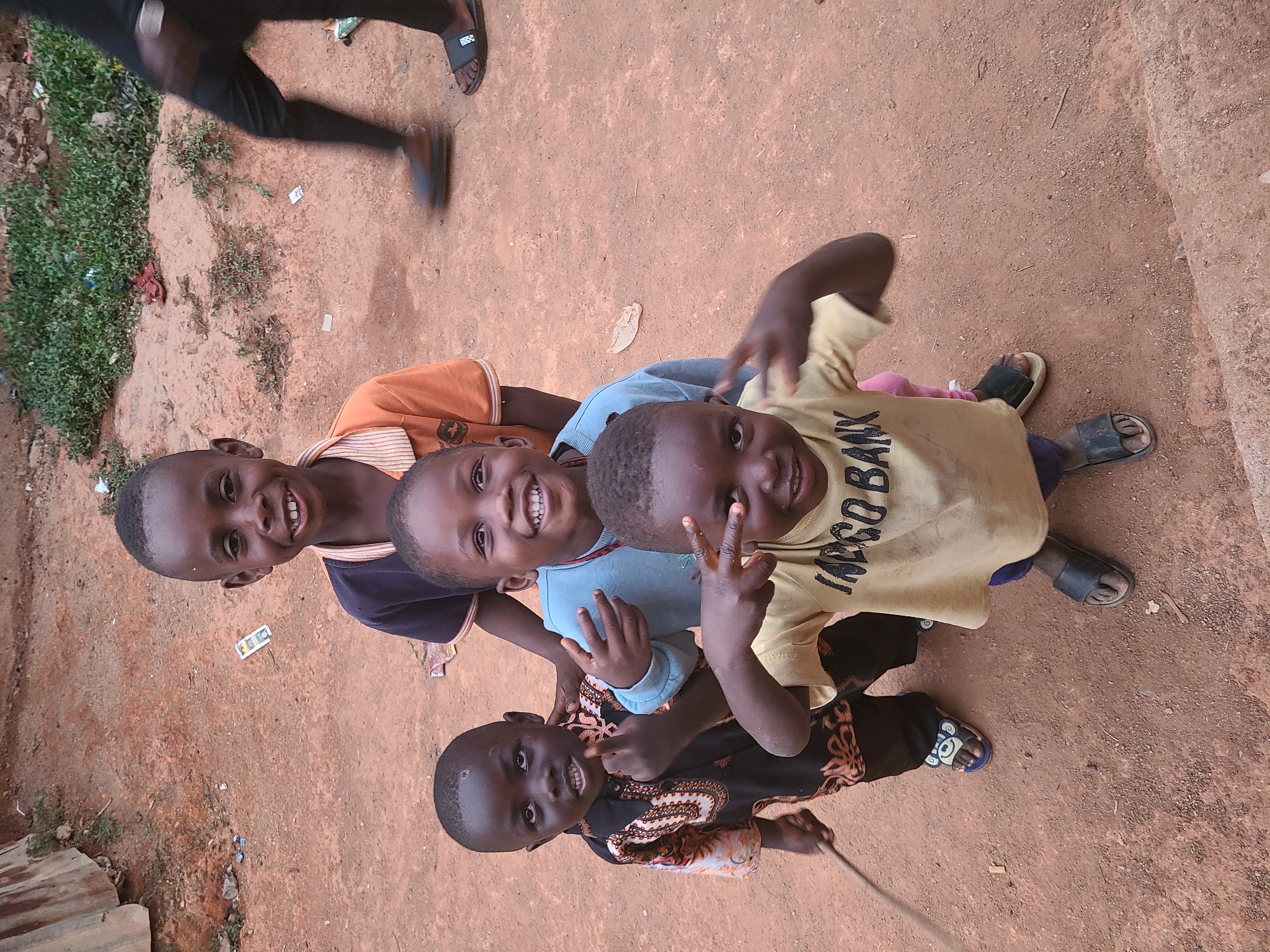 Four children are closely grouped together and looking up at the camera, all smiling. Three of the children are stacked vertically, and one is to the left. The top child is wearing a light yellow t-shirt with dark lettering, the second is in a light blue long-sleeve shirt, the third has an orange and navy striped shirt, and the bottom child is wearing a patterned brown and black shirt. A fifth person is seen partially in the top left corner, wearing dark clothes and sandals, apparently walking or running. The children are standing on a reddish-brown dirt surface. There is green vegetation along the left edge of the frame, and a corrugated metal edge is visible at the bottom left. The location is Jos, Nigeria.