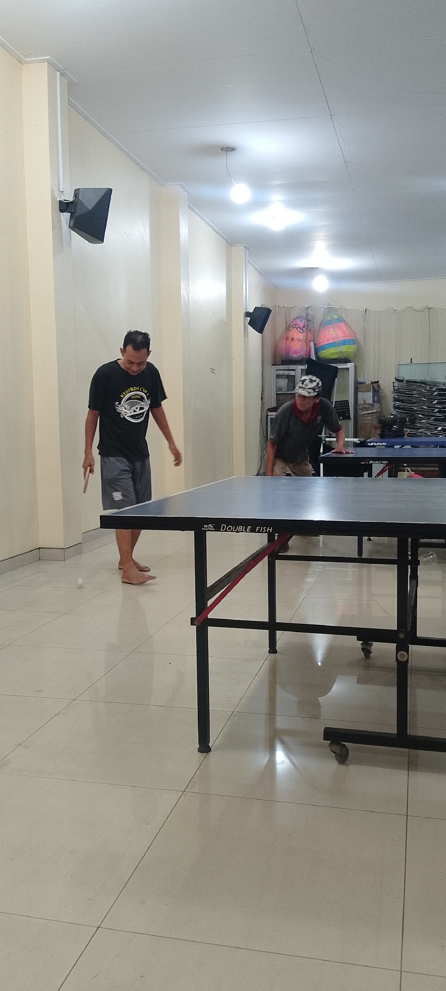 Two men are playing table tennis in a room in Kudus, Indonesia. The man on the left is wearing a black t-shirt and gray shorts, and is holding a table tennis paddle. He is barefoot and is looking down at the ball, which is in the air in front of him. The man on the right is wearing a gray t-shirt, khaki shorts, and a patterned cap. He is leaning over the table tennis table, which is the main object in the foreground. The room appears to be a recreational facility, with posters on the wall and other tables visible in the background. The floor is tiled and reflective, with a clear reflection of the ceiling lights. The walls are painted a light cream color.