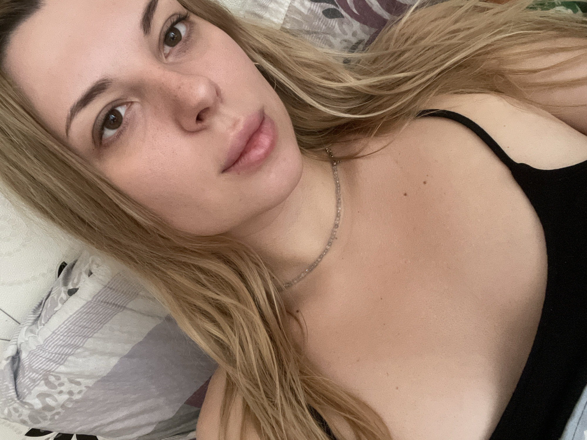 The image is a close-up selfie of a young Caucasian woman with long, blonde hair. She is lying down, possibly in a bed, as indicated by the patterned pillows and blanket in the background. The woman is wearing a black top and a delicate necklace. Her face is partially visible, with her eyes looking towards the camera. The lighting suggests an indoor setting. The image does not provide any information about the location in the city of Shcherbinka, Russia.