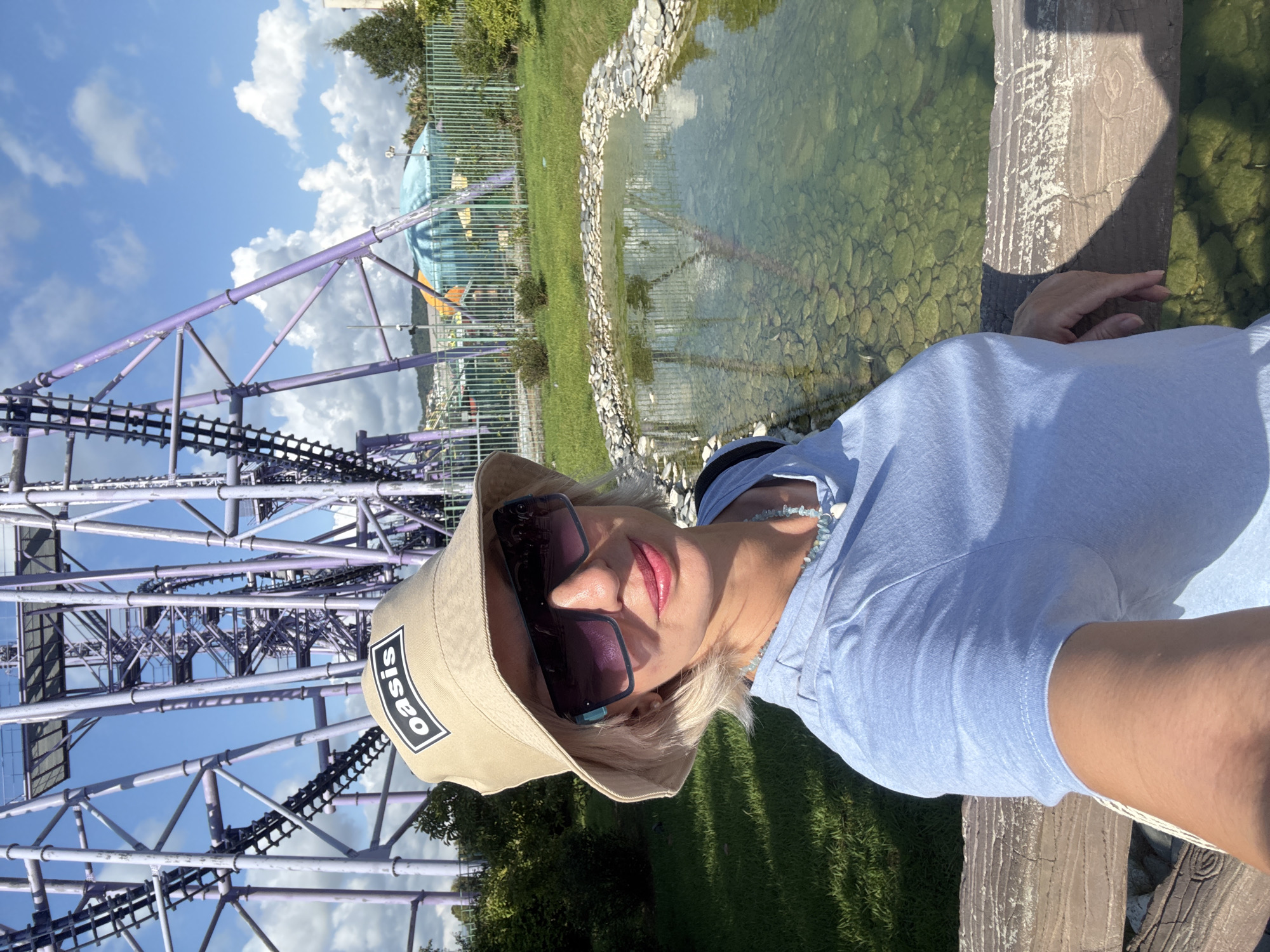 A woman with short blonde hair, wearing a tan bucket hat with an "oasis" logo, large dark sunglasses, a light blue t-shirt, and a beaded necklace, takes a selfie outdoors. She is smiling and leaning on a wooden railing. Behind her is a large purple roller coaster structure, a small decorative pond with stones, green grass, bushes, and in the distance, some colorful structures and a fence. The sky is blue with white clouds. The scene appears to be at an amusement park in Sochi, Russia.