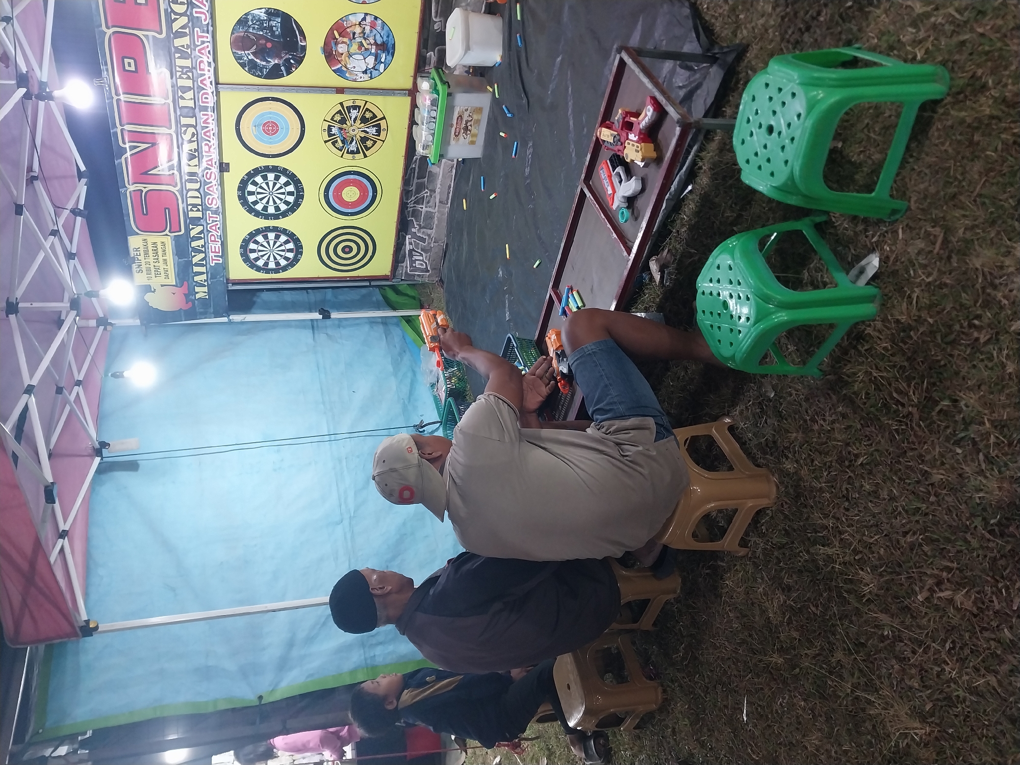 The image shows a carnival game stall in Jiwut, Indonesia. The stall features a "Sniper" themed target shooting game with various dartboards and toy dart guns. Three individuals, two men and a young boy, are seated on plastic stools in front of the game. One man is actively playing, aiming a dart gun at the targets. The ground is covered in grass, and a black tarp is laid out in front of the stall. Various accessories for the game, including darts and toy guns, are visible. The stall is covered by a colorful canopy, with bright lights illuminating the scene.