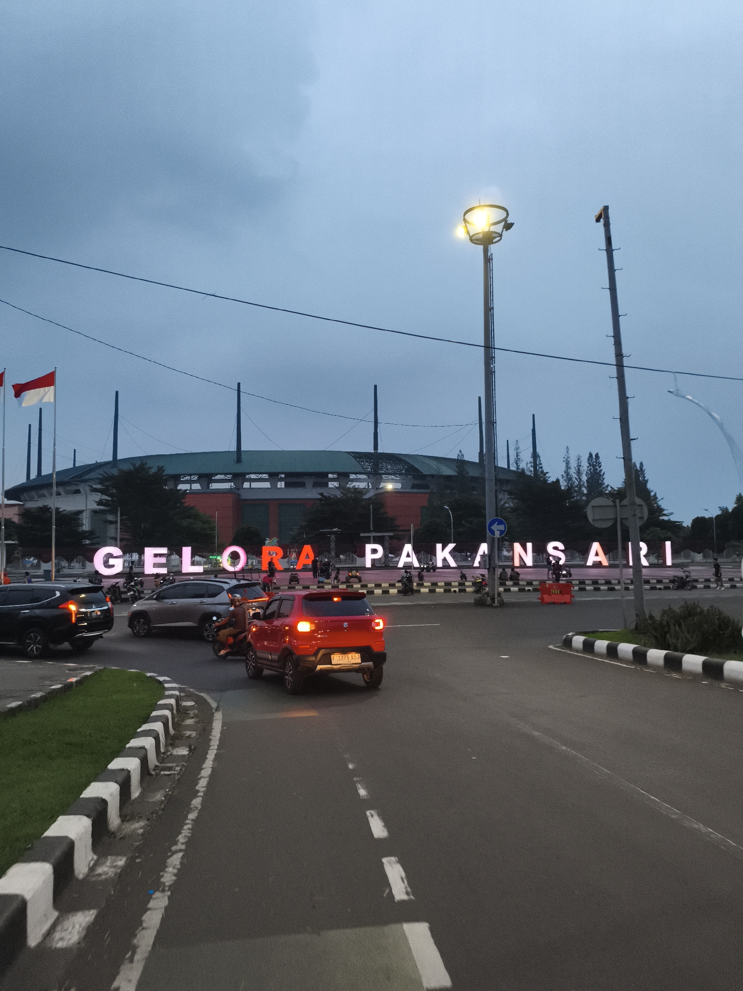 The image depicts an evening scene near the Gelora Pakansari Stadium in Cibinong, Indonesia. The sky is a muted, overcast blue-grey, indicating dusk or early night, with artificial lights starting to illuminate the area.
In the foreground, a paved road with white lane markings curves around a grassy median bordered by black and white striped curbs. Several vehicles are present: a black SUV on the left, a silver sedan in the middle, and a bright red compact SUV with illuminated taillights in the lower right. Multiple motorbikes are also seen, some moving and others parked, with riders and passengers visible.
The central focus is a large, illuminated sign spelling "GELORA PAKANSARI" in glowing letters. People are gathered around the base of this sign, suggesting a public area or entrance. Behind the sign, the stadium's architecture is visible, featuring a green roof and numerous vertical support structures. To the left of the stadium, two Indonesian flags are hoisted on flagpoles.
A tall streetlight with bright, glowing lamps stands prominently to the right of the stadium sign, while an unlit pole is next to it. Traffic signs, including a blue circular sign with a white arrow indicating a turn, and red-and-white traffic barriers, are also visible. The red SUV's license plate reads "F 3373 ABJ." The scene captures a typical evening in an urban environment, centered around a significant landmark.