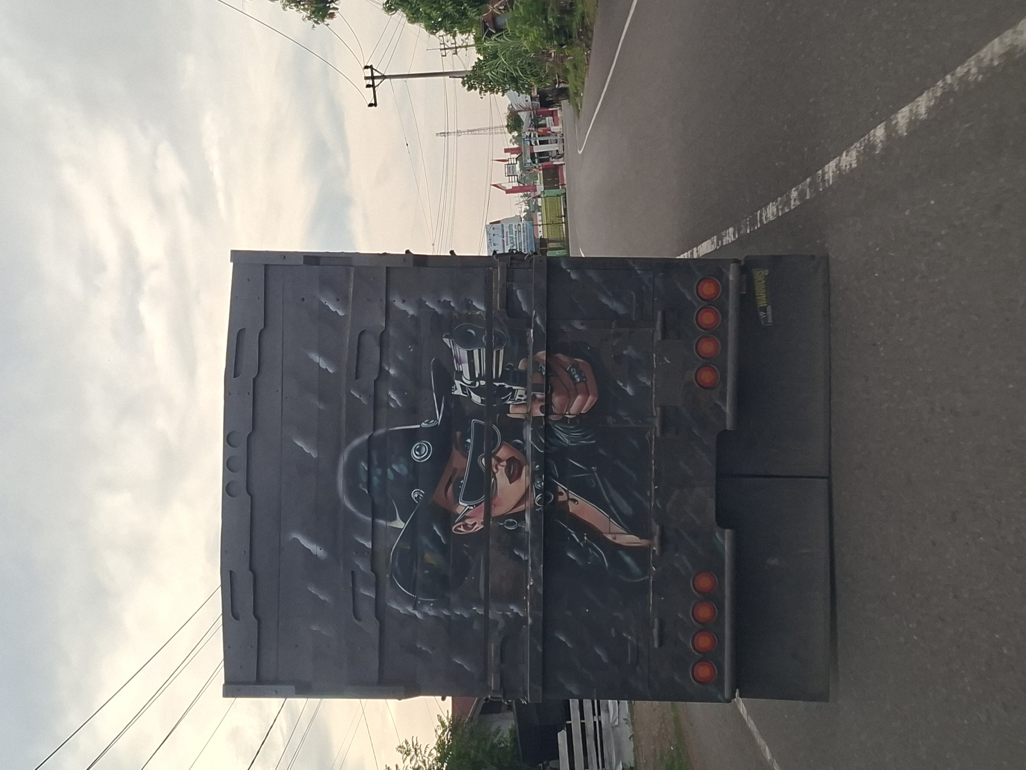The image shows the back of a flatbed truck driving on a paved road, likely within Mee Pangwa, Indonesia, based on the surrounding environment and context. The truck bed is painted with an image of a woman holding a revolver. Traffic lights are visible at the back of the truck. Overhead power lines run across the scene, and in the background, there are buildings and trees. The road surface is grey asphalt, with a visible white line marking the lane.