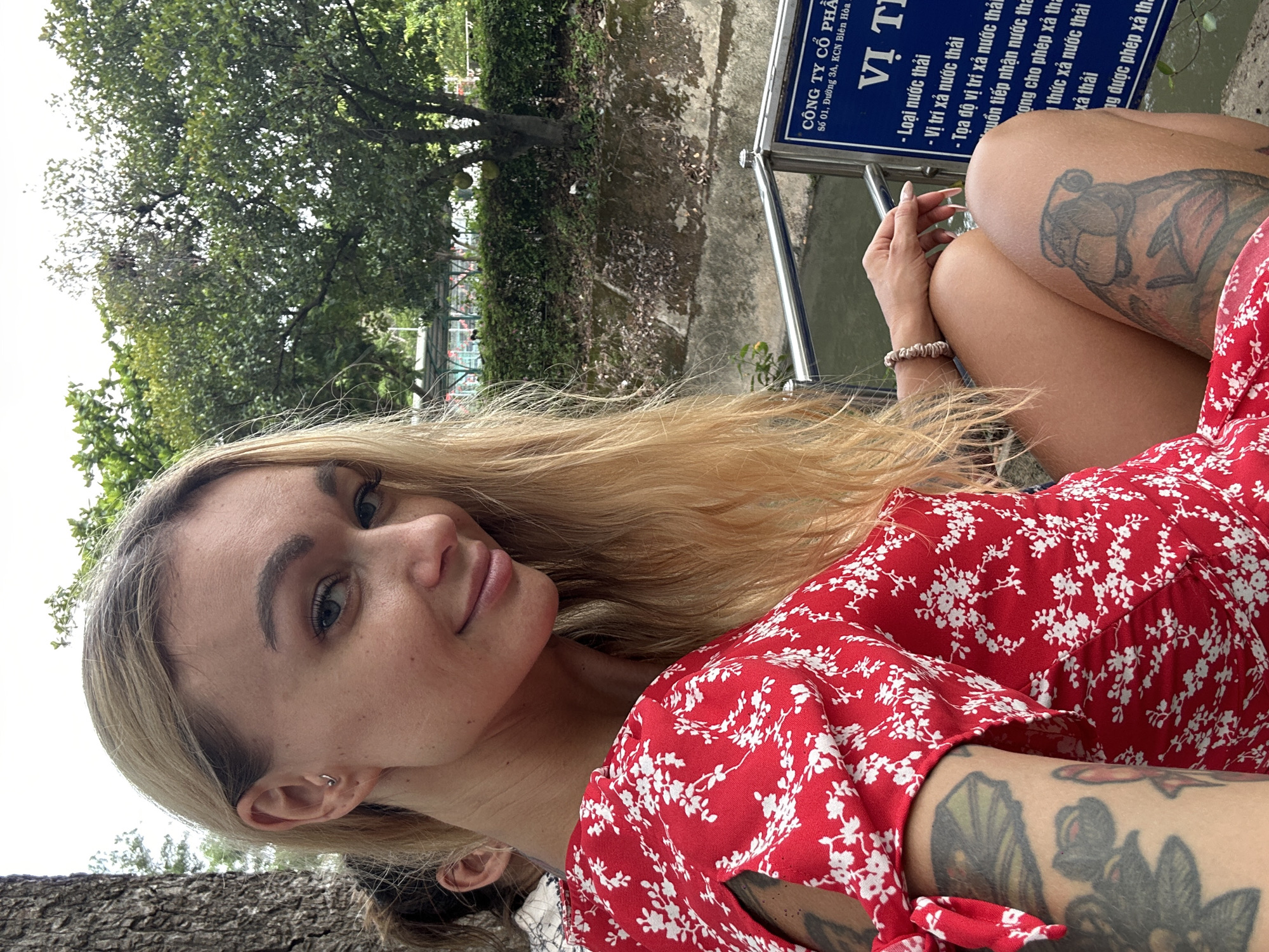 The image shows a woman with blond hair and tattoos, wearing a red floral dress, seated next to a blue sign. The sign contains Vietnamese text, which translates to "Công ty Cổ Phần" (Joint Stock Company) at "Số 01, Đường 3A, KCN Biên Hòa" (No. 01, Street 3A, Bien Hoa Industrial Zone). The title on the sign is "Vị Ti." The woman is sitting near a body of water.
