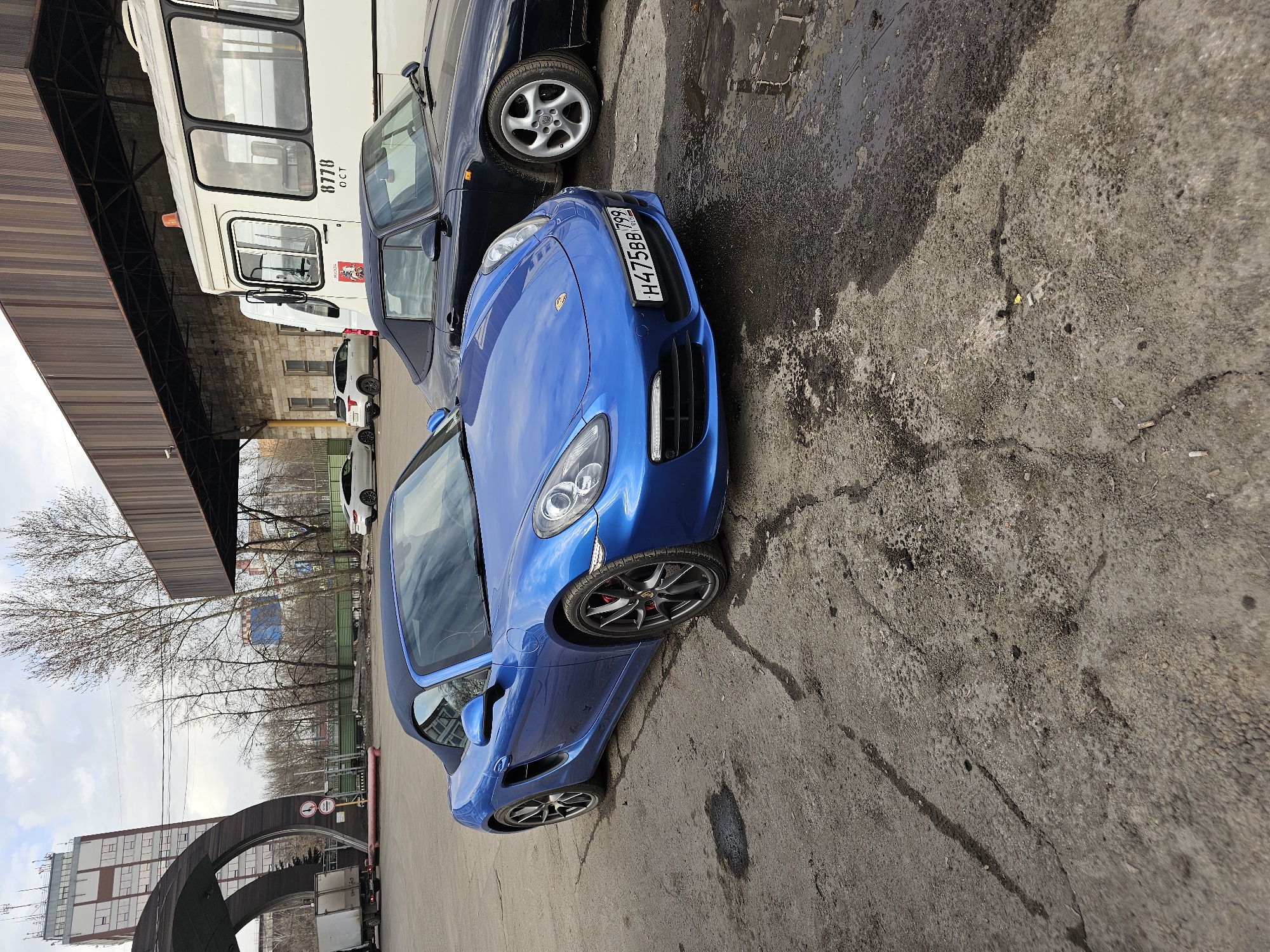 The image shows a blue Porsche parked on a concrete surface next to other vehicles, including a white bus or truck. The license plate of the Porsche reads "H475BB799". The location is likely Khimki, Russia, based on the Cyrillic characters on the license plate. The background includes bare trees, buildings, and an arched structure. The weather appears overcast.