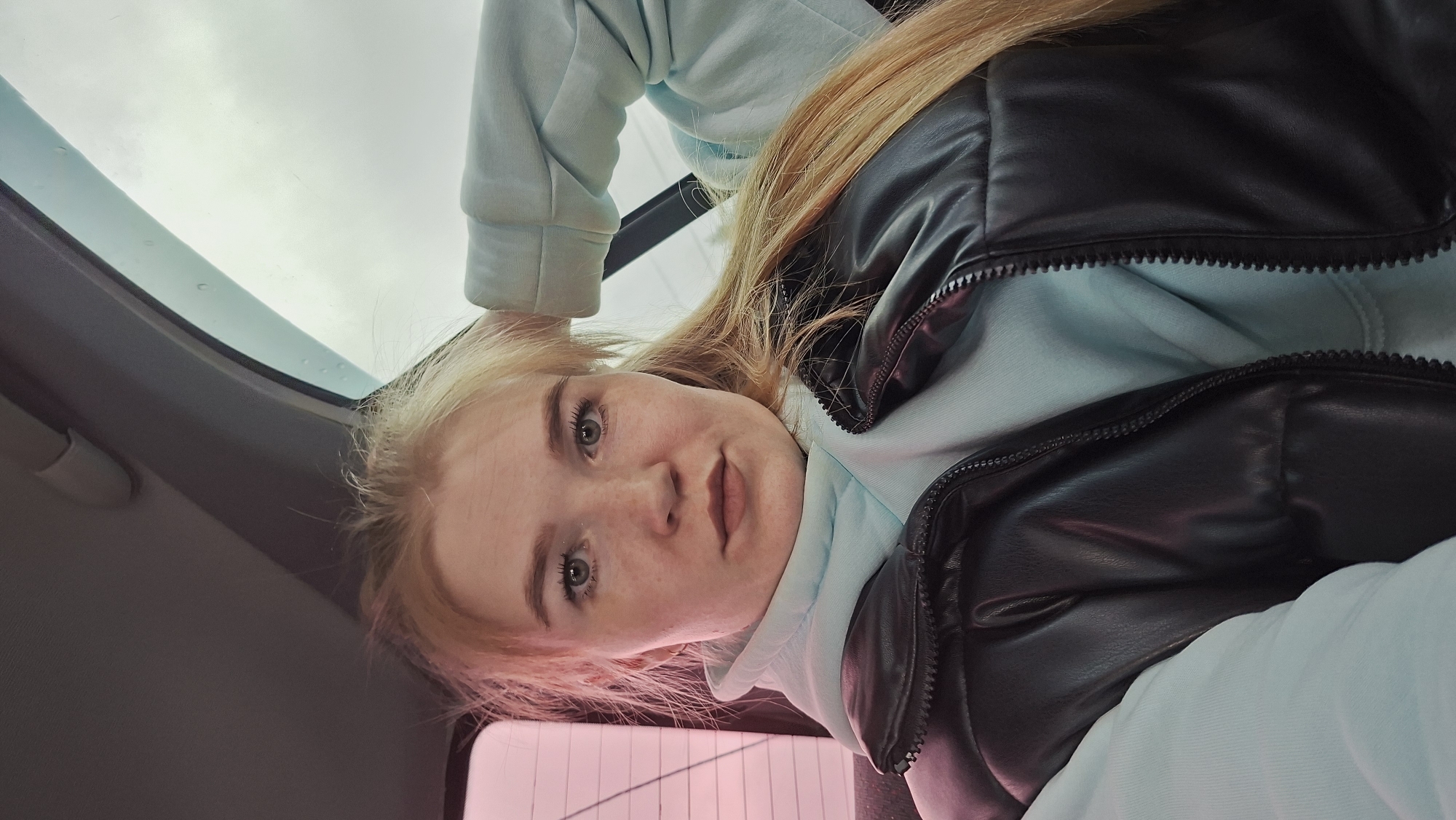 A woman with fair skin and light brown hair is looking directly at the camera from an unusual downward angle. Her expression is neutral, and she appears to be inside a vehicle. She is wearing a light blue hoodie with a high collar and a black vest. In the upper left corner of the frame, the edge of a car window can be seen, suggesting the scene is taking place inside the vehicle. The background is indistinct and out of focus, but a hint of pink or red is visible in the lower right, possibly indicating taillights or a similar light source. The sky is gray and overcast. The location is not identifiable.