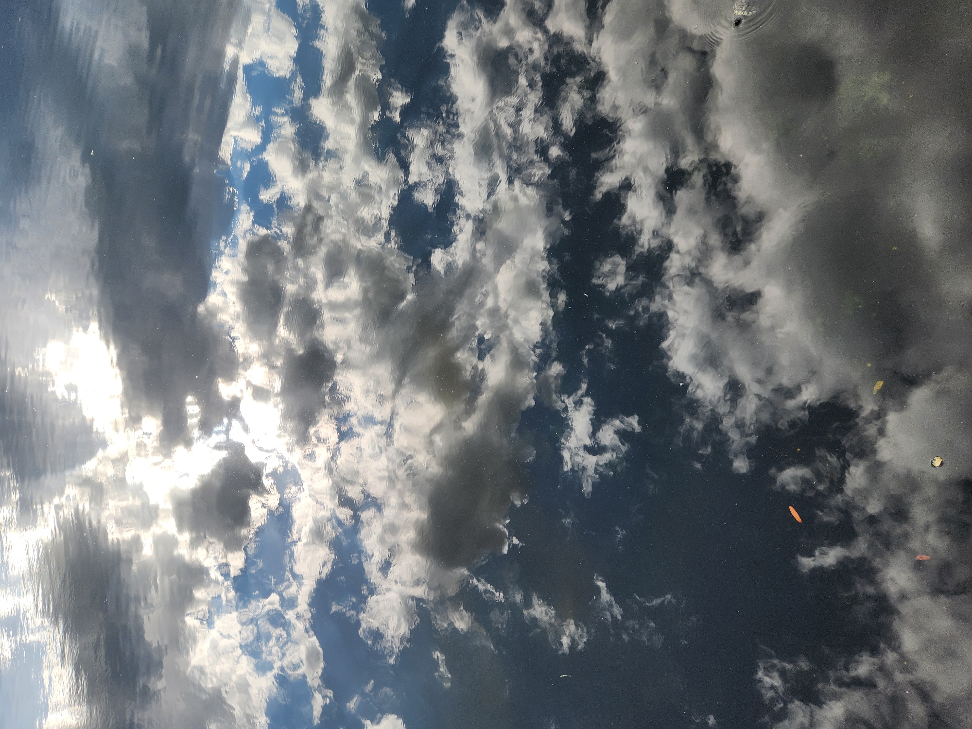 A close-up, high-angle, full-shot image captures the rippling surface of a body of water, reflecting a sky filled with dramatic clouds. The water's surface is dark, creating a stark contrast with the bright white and gray clouds that are distorted by the movement of the water. Patches of blue sky are also visible between the clouds, adding depth to the reflection. The overall impression is one of a dynamic and somewhat abstract natural scene. The location of this image, as part of the city of Maitland, United States of America, is noted, suggesting it might be a park, lake, or river within the city.