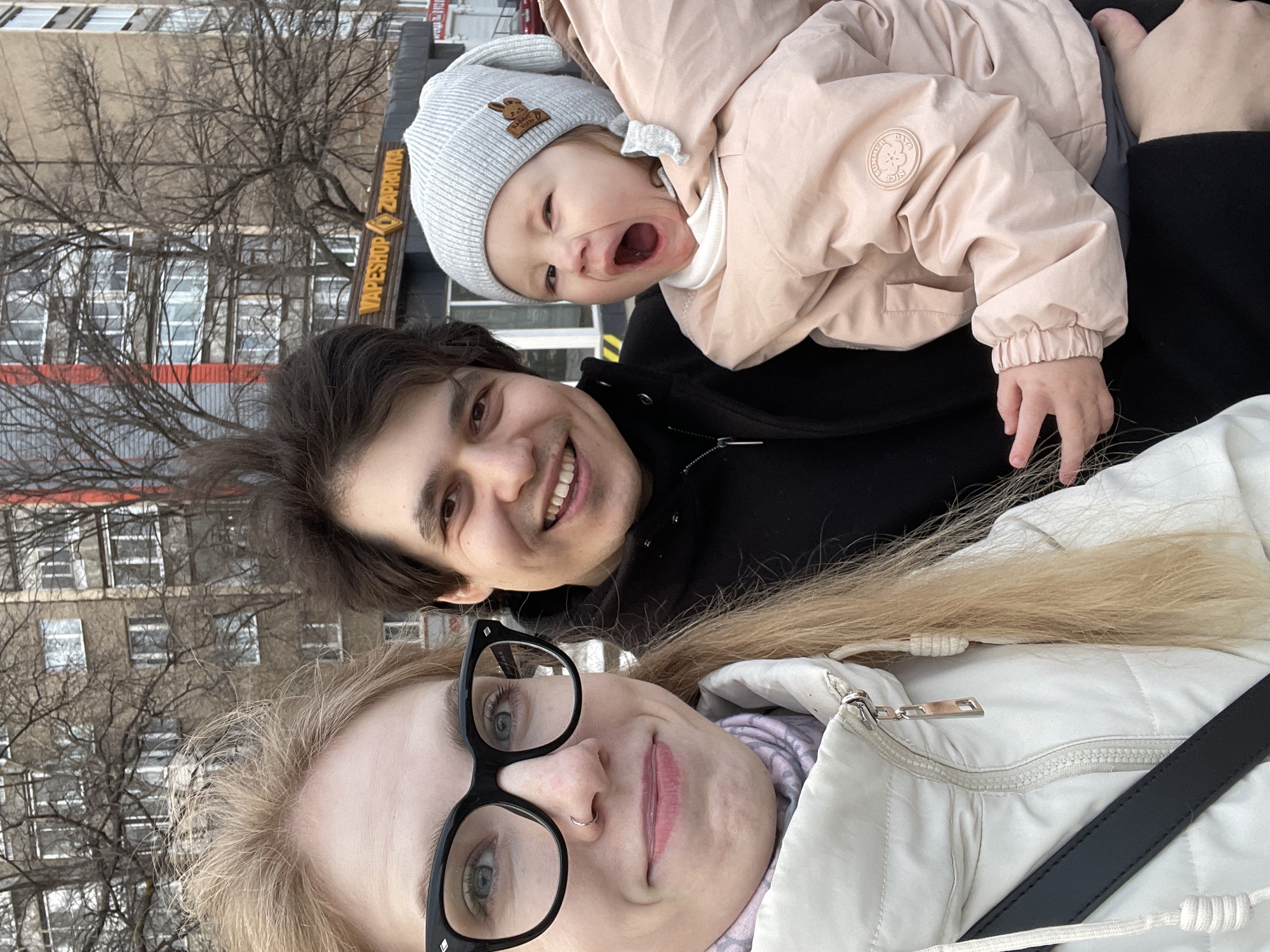 A family portrait of three individuals is captured in Izhevsk, Russia. A woman with blonde hair and black-rimmed glasses takes a selfie. Behind her, a man with dark hair smiles at the camera. An infant wearing a gray beanie with a rabbit design and a pink jacket is positioned above the man. The sign reading "Vape Shop Zapravka" is visible in the background. A multi-story building and bare tree branches fill the background. The overall setting is an outdoor, urban environment during the day.