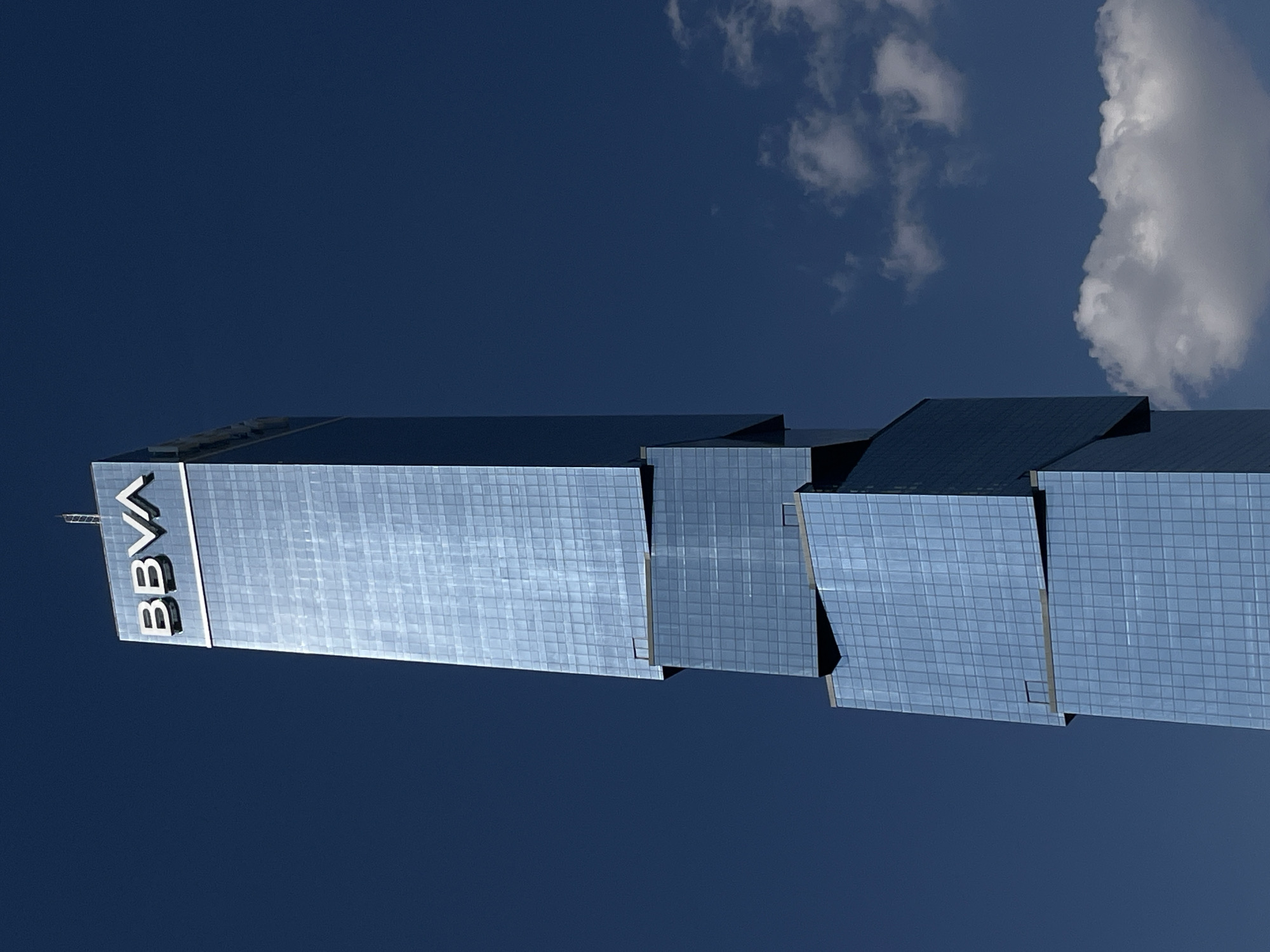 The image captures a low-angle view of the BBVA Tower in an unknown city, showing a portion of the glass building against a deep blue sky. The building is composed of several stepped sections. The BBVA logo is clearly visible on one of the sections. Two clouds are visible in the upper right corner of the frame. The overall tone is sharp and reflective, showcasing the modern architecture of the building.