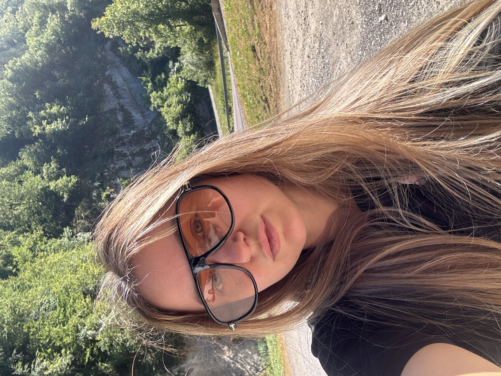 A young woman with long, blonde hair is posing for a selfie outdoors. She is wearing large, dark-rimmed sunglasses with orange lenses and a black t-shirt. The background shows a lush, green, forested hillside with a rocky cliff face and a road winding through it. The ground in the foreground is a gravelly dirt path. The lighting suggests it is a sunny day. There is no indication that this location is in L'Essard, France.