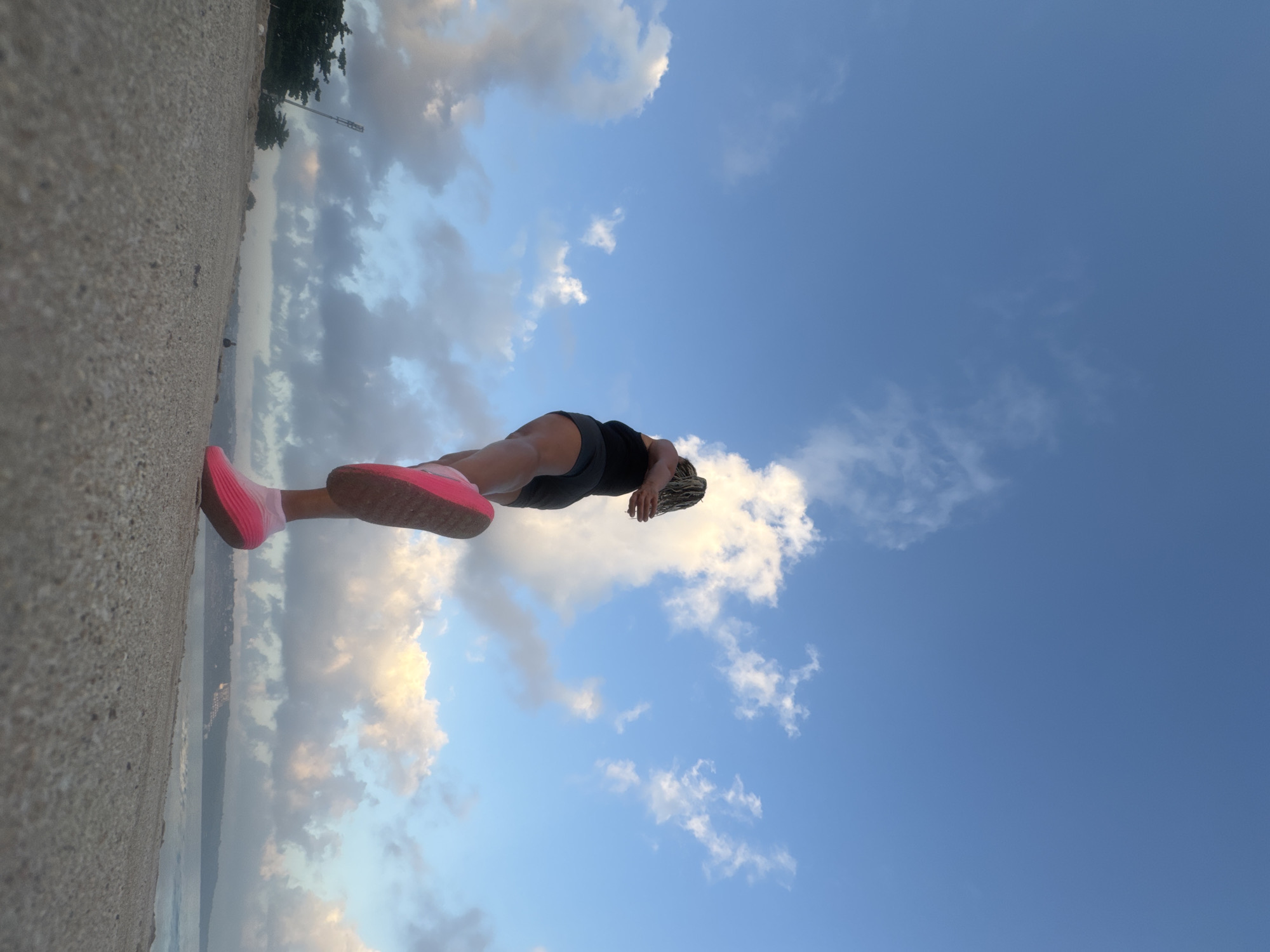 The image shows a low-angle, wide shot of a person running on a sandy beach in Kuta Selatan, Indonesia. The person is wearing black shorts and a black tank top, with bright pink running shoes and socks. Their hair is in blonde braids. The person is positioned in the lower right quadrant of the frame, with their back to the camera. The beach sand occupies the foreground, with a clear view of the ocean and a distant coastline in the background. The sky above is a vibrant blue, with scattered white and grey clouds, suggesting it is either early morning or late afternoon. A single streetlight can be seen in the distance on the left side of the image, near some trees. The overall scene depicts an outdoor activity in a coastal environment.