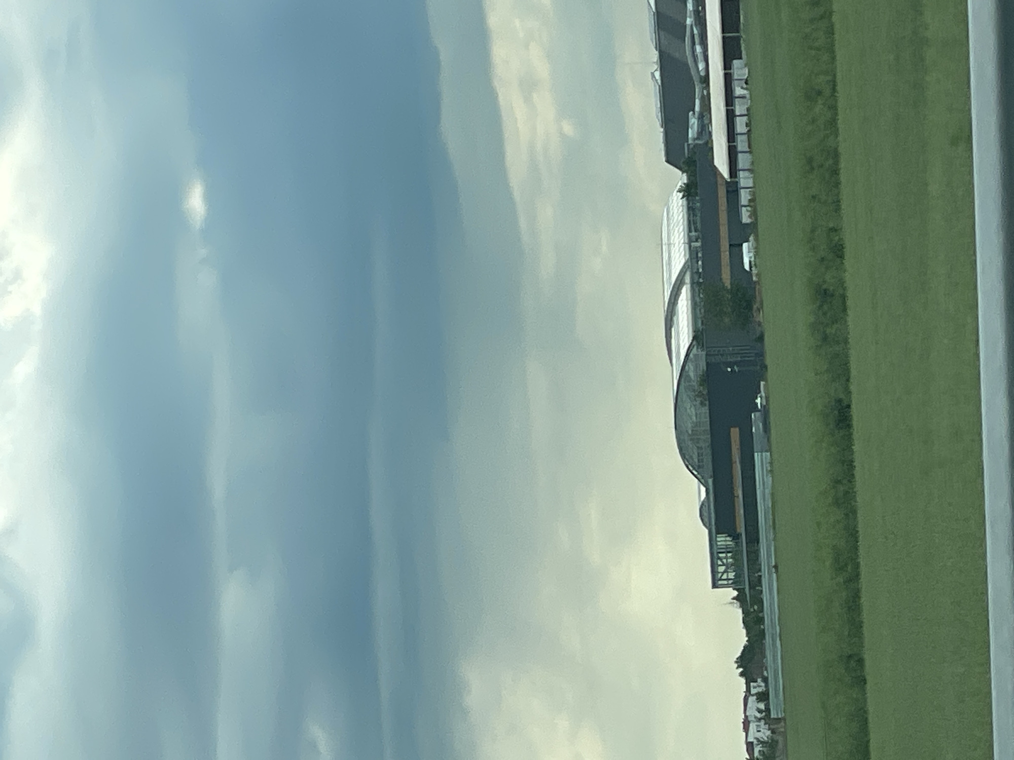 The image shows a building complex, possibly a sports stadium or event venue, in Balotești, Romania. The building has a curved, glass-covered section and a more rectangular, multi-story section. The complex is bordered by a green grassy field. The sky is overcast with clouds.