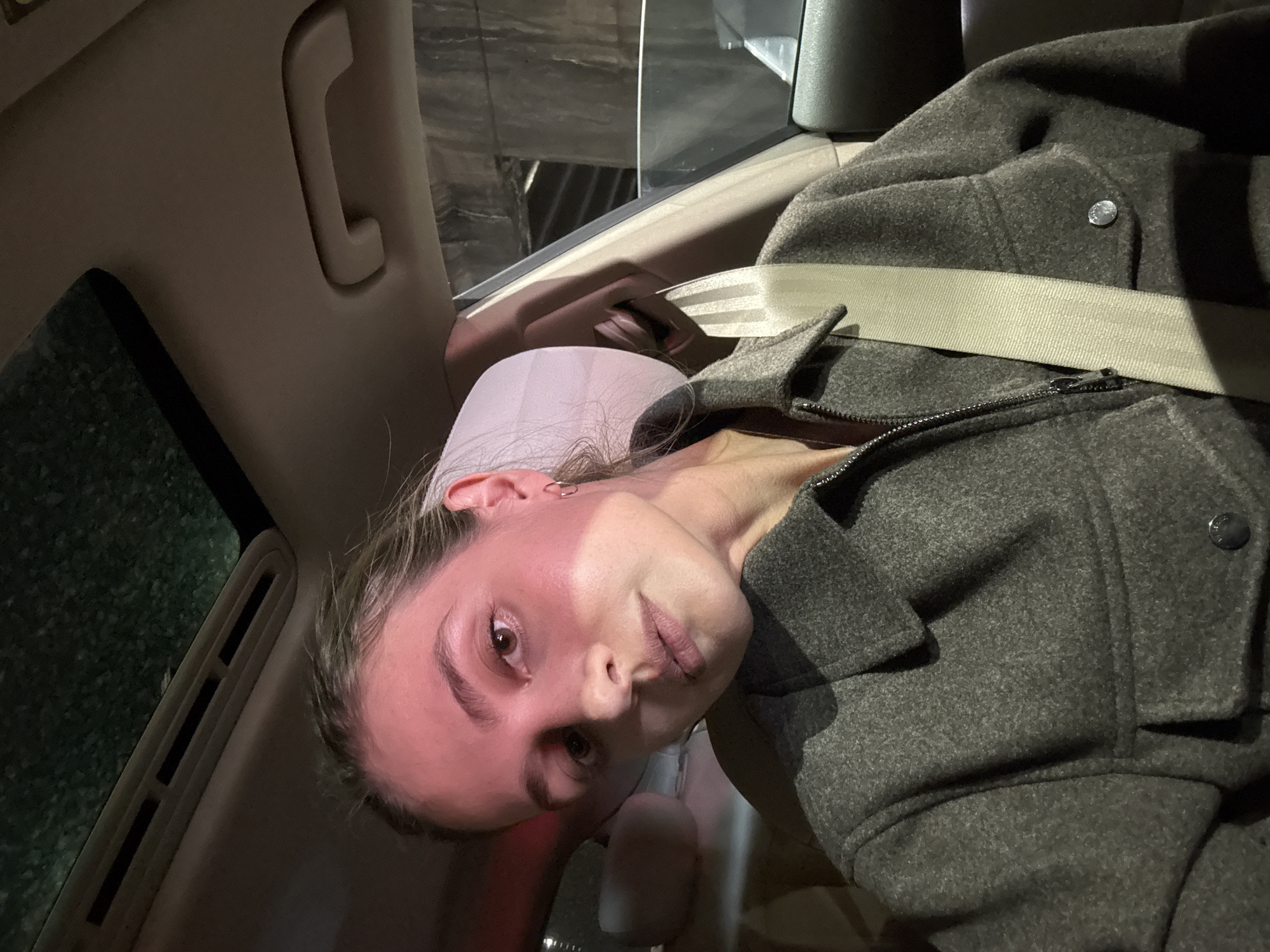 A young woman is seated in the back seat of a car, wearing a gray wool coat and a seatbelt. The car is moving at night, with outside lights visible through the window. The interior of the car is visible, including the headrest and the grab handle. The location appears to be Tbilisi, Georgia, based on the architecture visible outside the window.