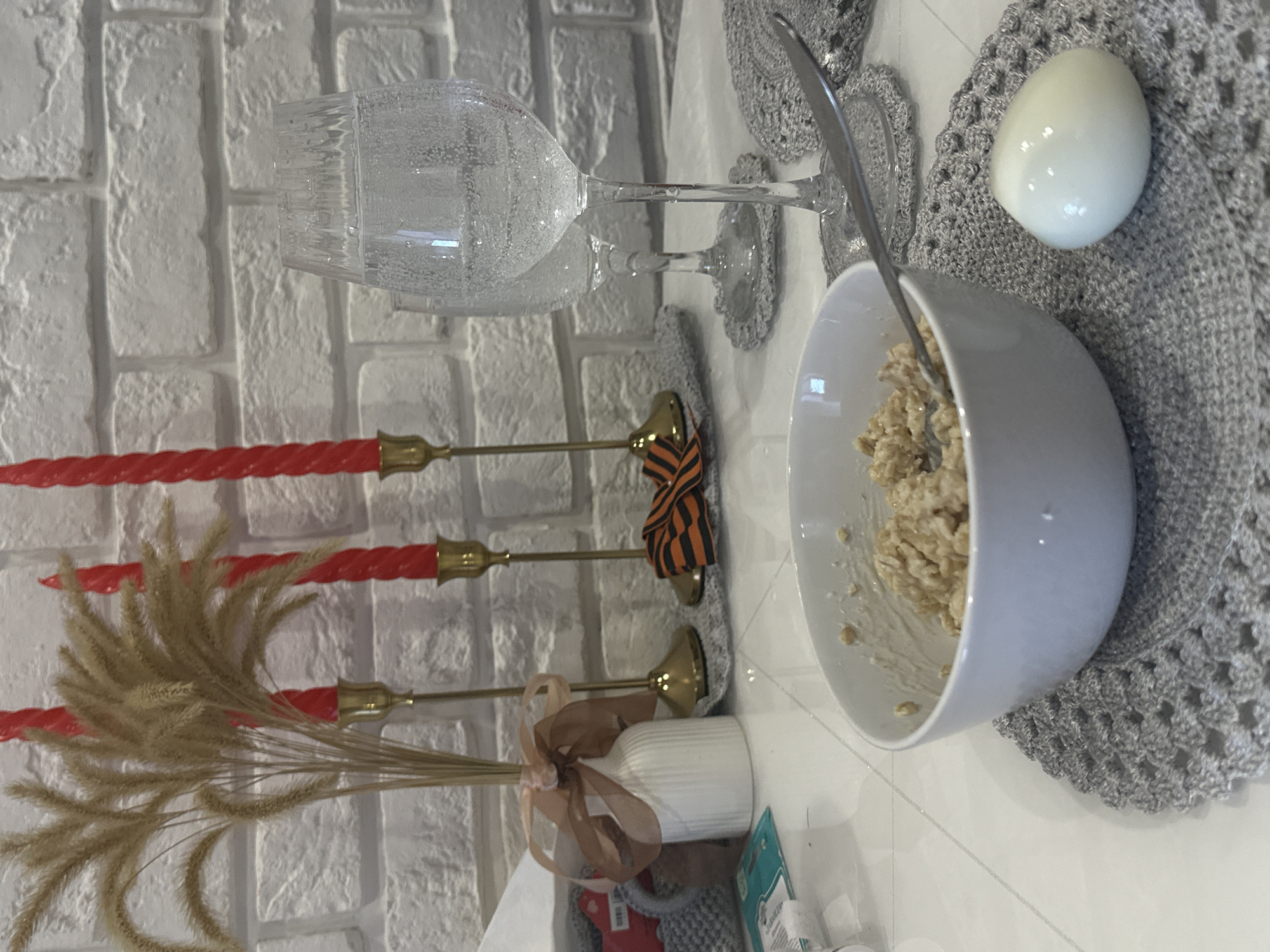 A still life is presented with a variety of items arranged on a white surface, likely a table. In the background, a brick wall is visible. From left to right, a vase with tall, dried plants tied with a light brown ribbon, three candelabras with red candles, a clear glass with liquid, a small, striped orange and black ribbon tied around a candlestick, a silver spoon, a bowl of oatmeal, and a hard-boiled egg on a gray crocheted doily. The location is not specified.