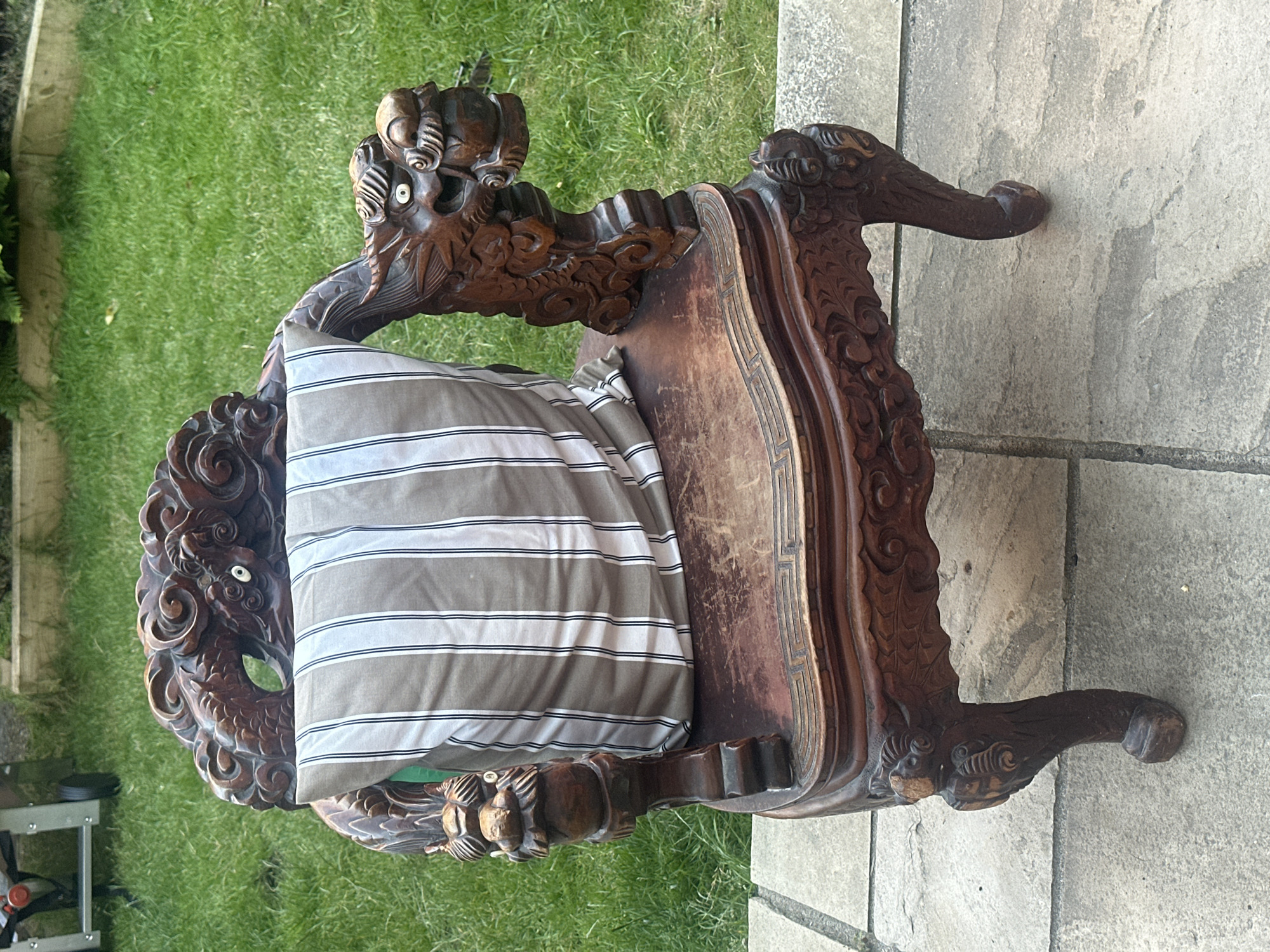An intricately carved dark wood chair, possibly antique, with dragon motifs on the armrests and back, is placed on a paved patio area outdoors. A grey and white striped cushion is propped against the back of the chair. The background consists of a well-maintained green lawn and a wooden garden border. The setting appears to be a domestic garden in Folkestone and Hythe District, United Kingdom. There are no people or animals visible in the image.