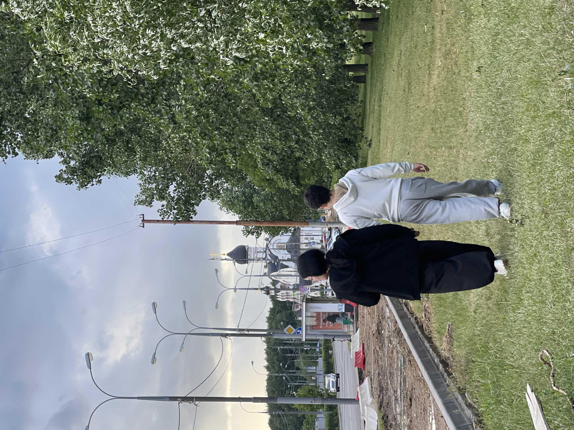 Two individuals, viewed from behind, walk across a grassy verge. The person on the left wears a black hooded jacket and loose black pants, with white sneakers. The person on the right wears a light grey hooded sweatshirt and light grey sweatpants, also with white sneakers. They appear to be walking together, holding hands. In the background, a road is visible with several cars. A bus stop with signage in Cyrillic script stands adjacent to the road. Further in the background, a white church with black domes and a golden cross is prominent. Large green trees line the right side of the frame, with a cloudy sky overhead. The scene is set in a suburban area of Moscow, Russia, with construction barriers visible on the left side of the road.