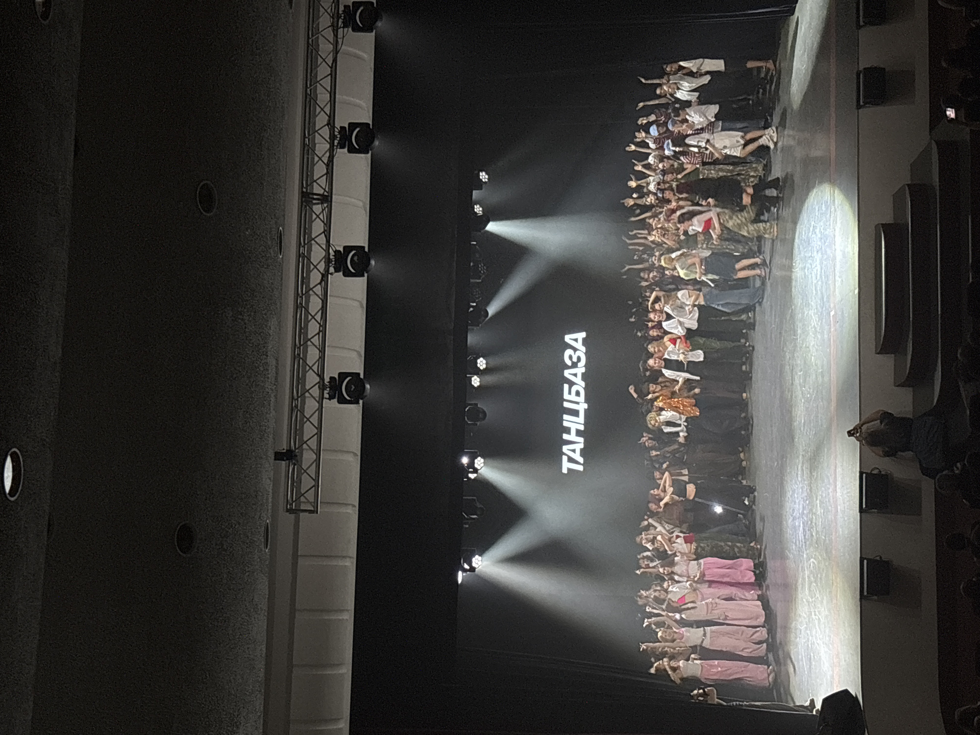 The image shows a stage performance in a theater, likely located in Perm, Russia, judging by the text on the screen "ТАНЦБАЗА," which translates to "Dance Base." A large group of people is assembled on the stage, possibly performers taking a bow or posing at the end of a show. Stage lighting is prominent, with several spotlights focused on the group. A lighting rig is visible to the left of the stage. The photograph is taken from an audience perspective, showing the performance and some elements of the theater's interior. A person holding a phone is visible at the right of the stage, presumably taking a photograph or video of the performance.