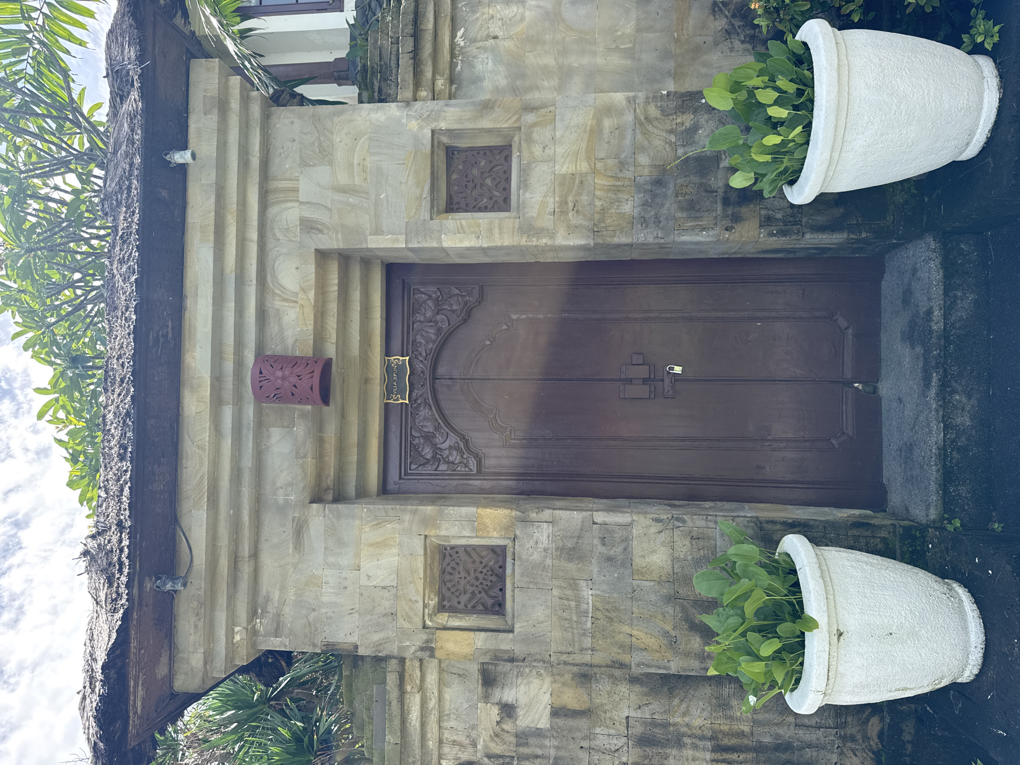 The image captures the entrance to a building, likely a villa, featuring traditional Balinese architecture. A grand, dark brown wooden double door with intricate carved details on its upper panels stands centrally. A small, golden rectangular plaque is affixed to the left door, faintly displaying the text "VILLA JEPUN."
The door is set into a wall constructed from light-colored, rectangular cut stone blocks, which exhibit natural variations in tone and some weathering. Two square recessed panels, positioned above and below the door, each contain a dark, ornate lattice or carved design. To the left of the door, a reddish-brown ceramic wall sconce, adorned with a floral pattern, is mounted on the stone.
Flanking the entrance are two large, white, textured ceramic planters, each holding vibrant green leafy plants. Above the doorway, a thick, dark wooden beam supports a substantial thatched roof, characteristic of tropical dwellings. A white security camera is discreetly mounted on this wooden beam on the left side.
Lush green tropical foliage, including palm fronds, is visible in the upper left of the frame against a bright, cloudy sky, indicating an outdoor, daytime setting. The ground in front of the door is composed of darker stone or concrete. The overall scene suggests a serene, traditional, and well-maintained property in a tropical environment.