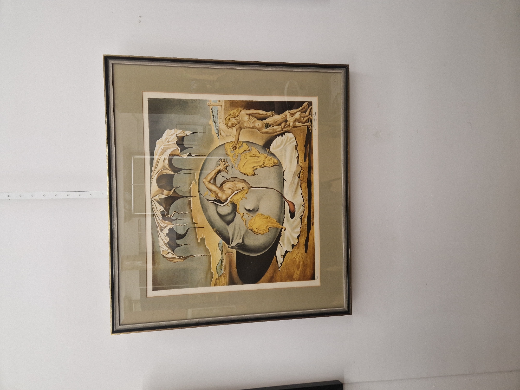 A framed print hangs on a white wall. The print depicts a surreal scene, possibly a reproduction of a work by Salvador Dalí, with figures floating around a central sphere with faces emerging from it. The frame is dark with a gold trim. The lighting is ambient and the surroundings are indistinct.