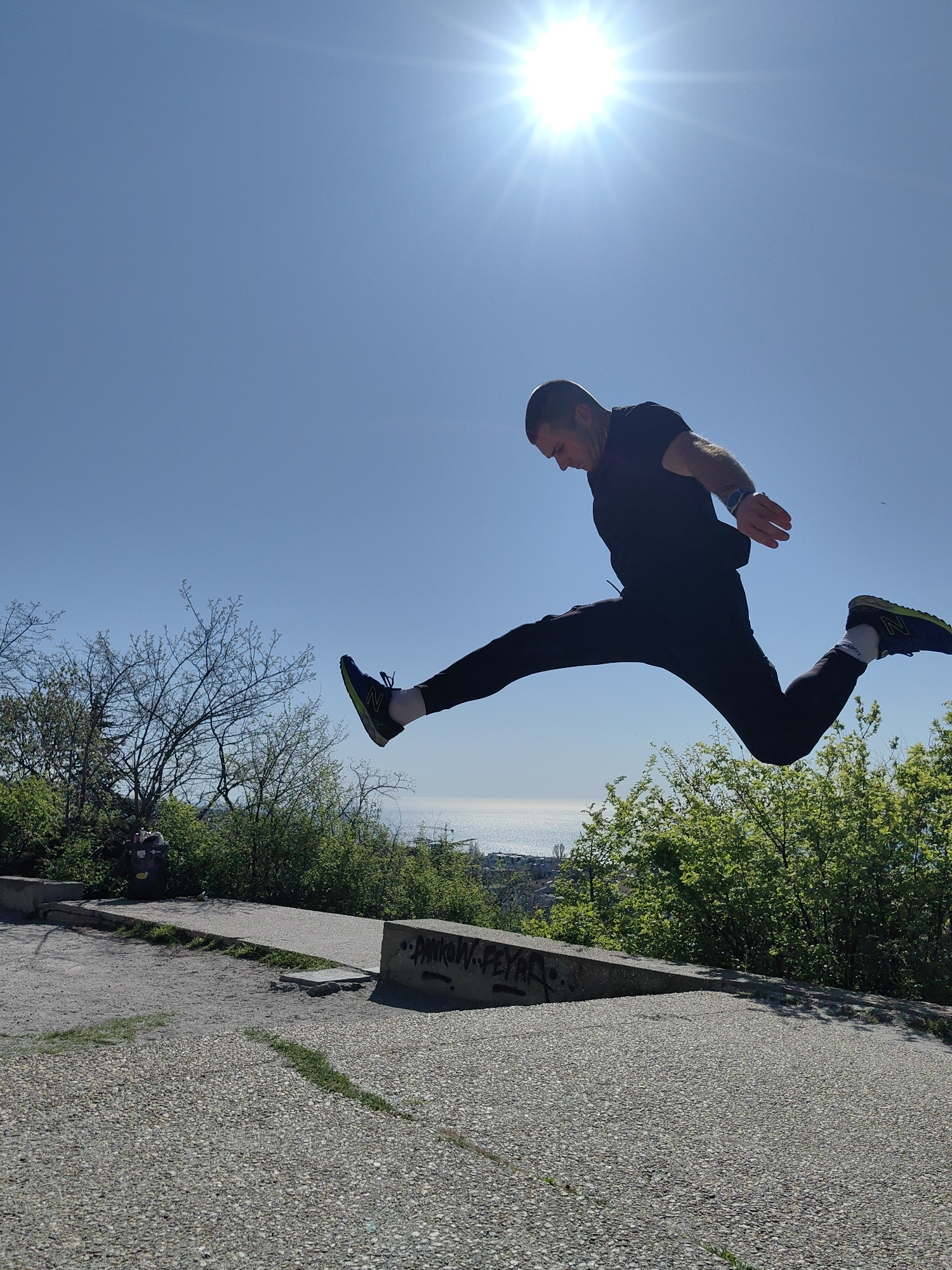 A man in athletic attire is captured mid-air in a jumping split against a backdrop of a bright blue sky and sunlight. He is wearing a black t-shirt, black athletic pants, and running shoes with white socks. He appears to be jumping over a small, concrete structure with graffiti on its side. Behind the structure is lush green vegetation and a glimpse of the sea in the distance. The foreground consists of a concrete surface with a pebbled texture. The location appears to be a park or public area with natural elements and urban features. The city is Unknown, Unknown.