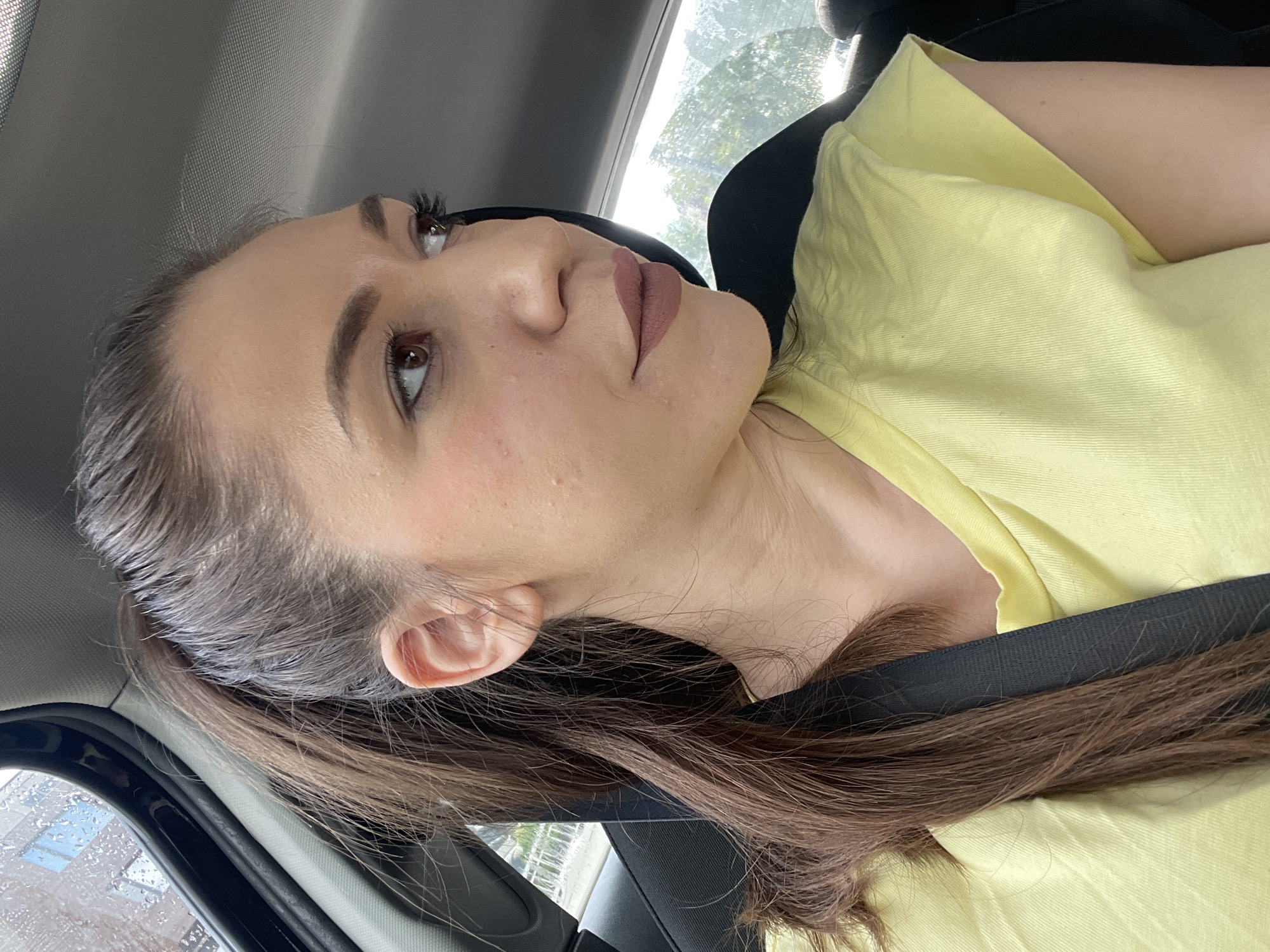 A close-up, profile-oriented shot features a woman in what appears to be the passenger seat of a car. Her face, turned upwards and to her left, shows a neutral to contemplative expression. She has dark brown hair, styled away from her face, and brown eyes with defined eyebrows and visible eyelashes. Her lips are adorned with light brown or nude lipstick.
She is wearing a light yellow t-shirt or top, and a black car seatbelt crosses her chest. The car's interior, including a dark headliner and seat, is visible around her. Through a window on the left side of the frame, raindrops are visible, suggesting rain or recent rainfall outside. The scene is brightly lit by natural light, indicating daytime. No text is visible in the image. The general location is Sofia, Bulgaria.