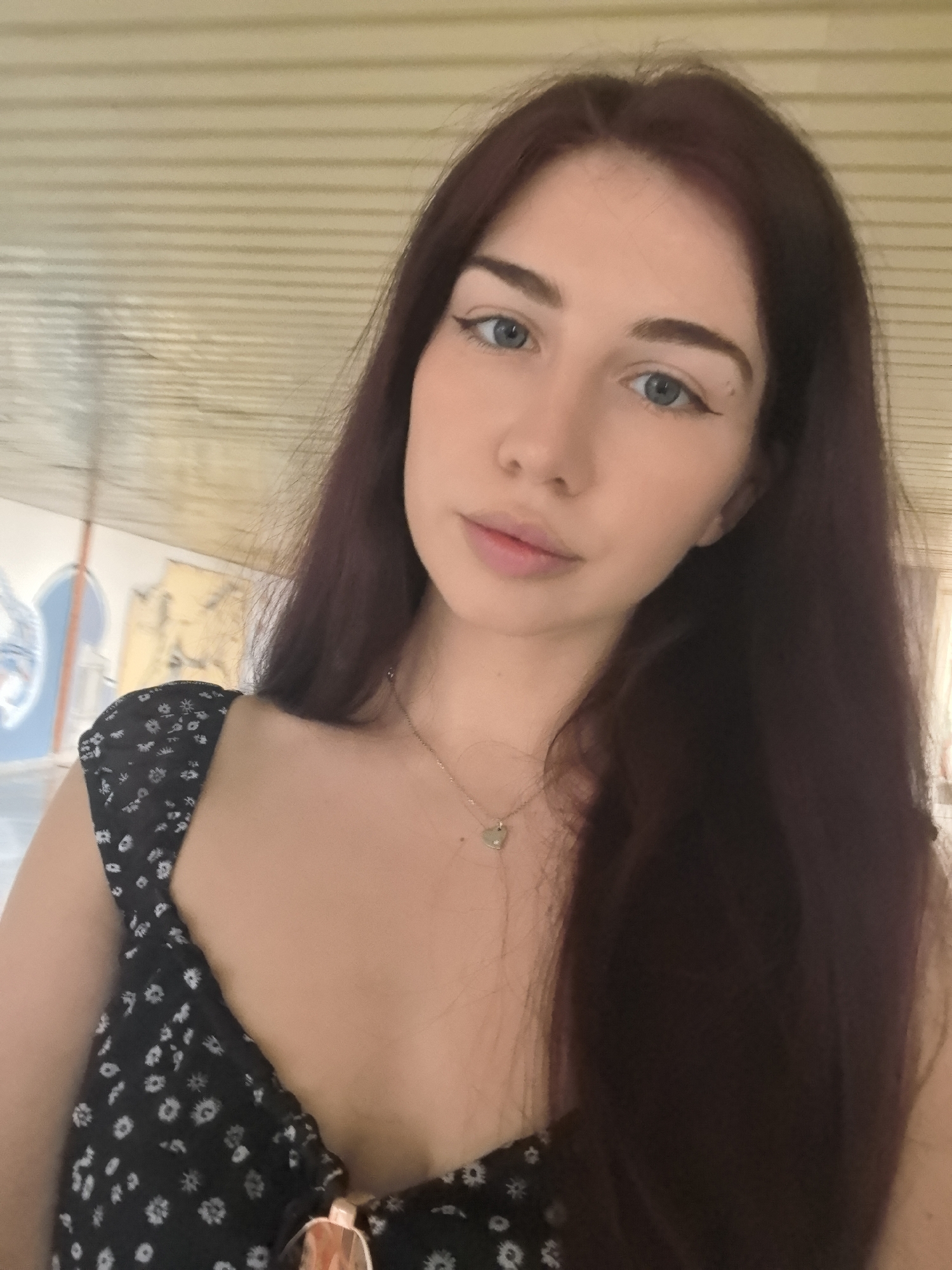 The image shows a young woman posing for a selfie, looking directly into the camera. She has long, dark reddish-brown hair, parted in the middle, and striking light blue-grey eyes accentuated with winged eyeliner. Her lips are a natural pink shade, slightly parted. She is wearing a dark-colored top, likely a dress or camisole, with a small white floral or dotted pattern. A delicate silver necklace with a heart-shaped pendant hangs around her neck, and sunglasses are visible clipped to her top in the lower right corner.
The background suggests an indoor or covered outdoor setting in Stara Zagora, Bulgaria. Above her head, a light-colored surface with horizontal grooves or panels is visible. To her left, there are hints of architectural elements, possibly arches or decorative structures, in light blue and white, suggesting a unique building or public art space. The lighting is soft and even, indicating daytime. Her expression is calm and neutral. There is no visible text in the image.