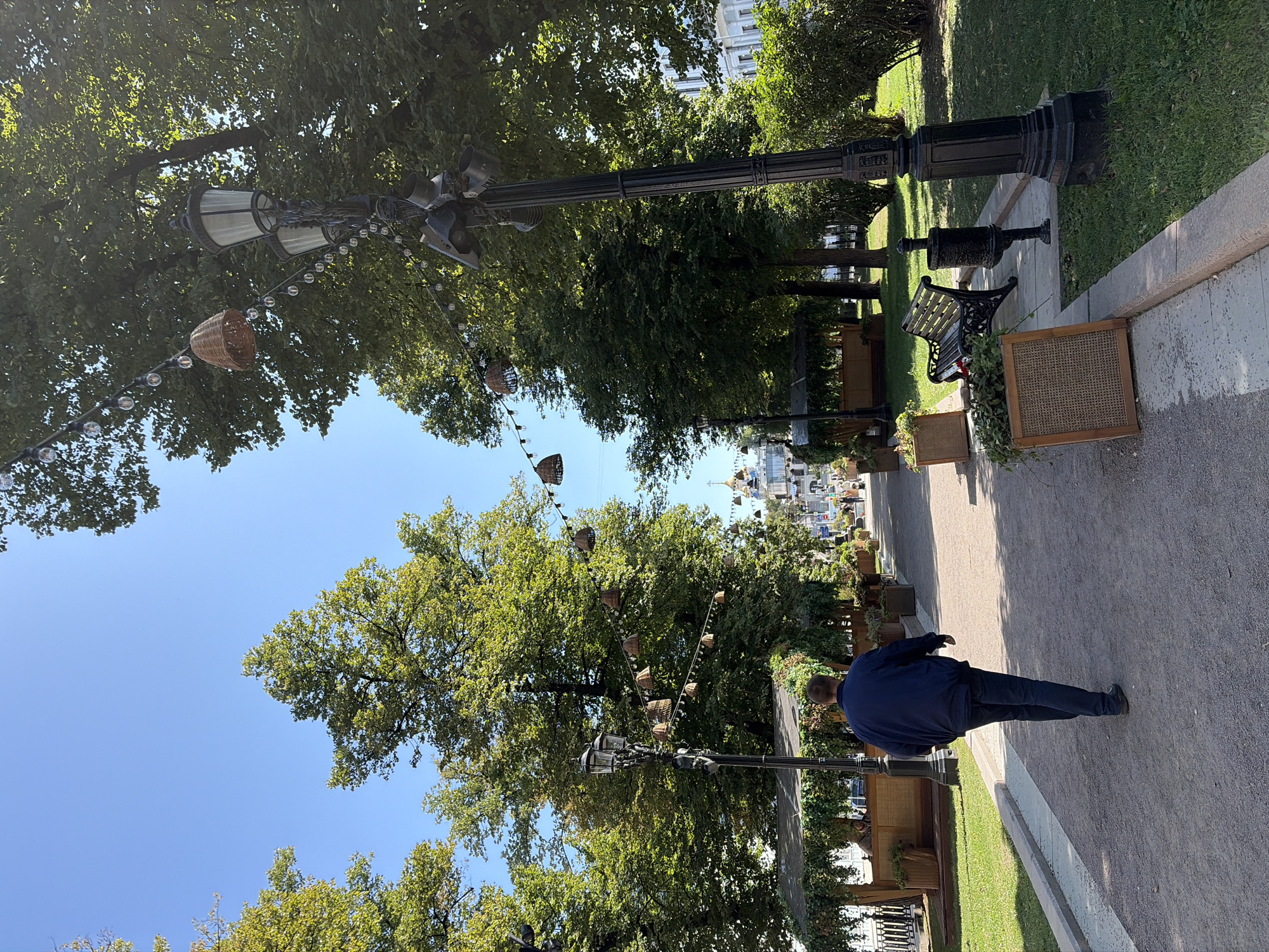 The image depicts a sunny day in a park-like setting, possibly a pedestrian area in Moscow, Russia. A man in a dark blue jacket and jeans walks away from the camera on a gravel path. To his left is a grassy area with trees, a covered seating area, and planters. To his right is another grassy area with a black cast-iron bench, a trash receptacle, and a large black lamppost adorned with hanging lights and woven basket decorations. Further down the path, more lampposts, trees, and buildings with golden domes are visible in the distance. The sky is clear blue.