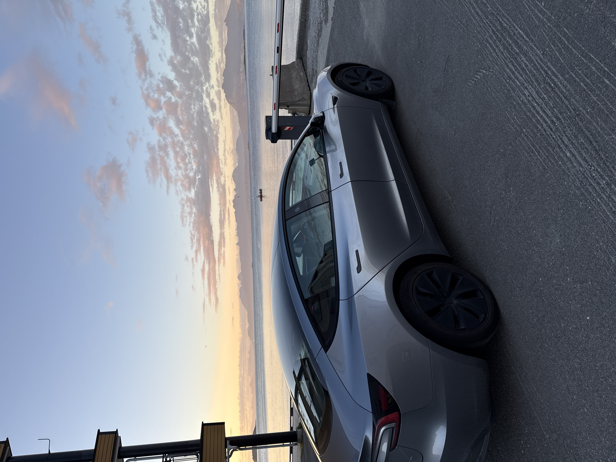 A silver Tesla Model 3 is parked on a paved surface next to a body of water. The car is facing towards the right of the frame, with its rear end closer to the viewer. Behind the car, a barrier gate is partially lowered across a roadway. In the distance, a calm body of water is visible, with mountains on the horizon. The sky above is a soft gradient of blue, with wispy clouds tinged with pink and orange from what appears to be a sunrise or sunset. To the left of the car, a portion of a dark building with wooden accents is visible. The scene is likely set in Harstad, Norway, given the coastal landscape and architecture. There are no people or animals visible in the image.