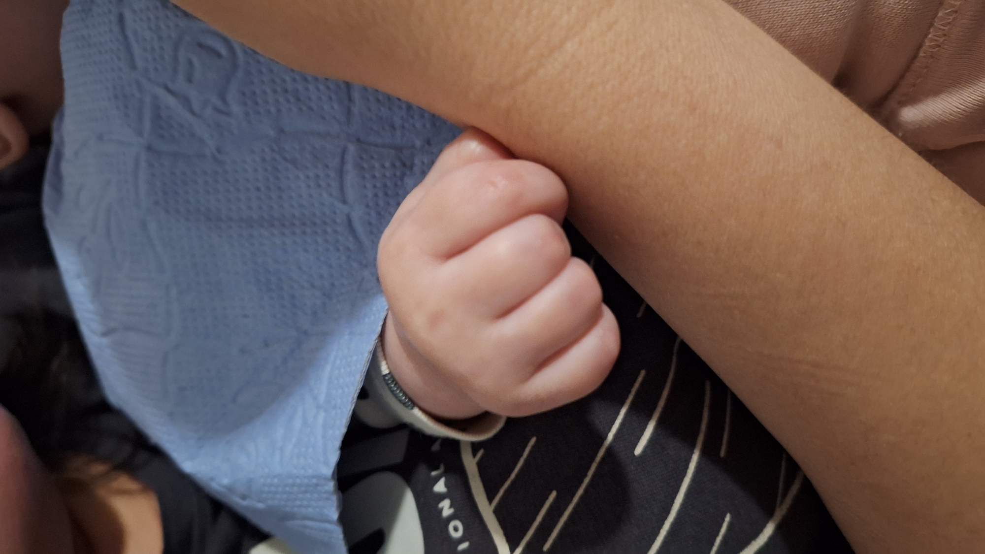 A baby's hand is tightly clenched, held against the arm of an adult. The baby is wrapped in a blue blanket. The background includes a dark fabric with white stripes. The location is not discernible from the image.