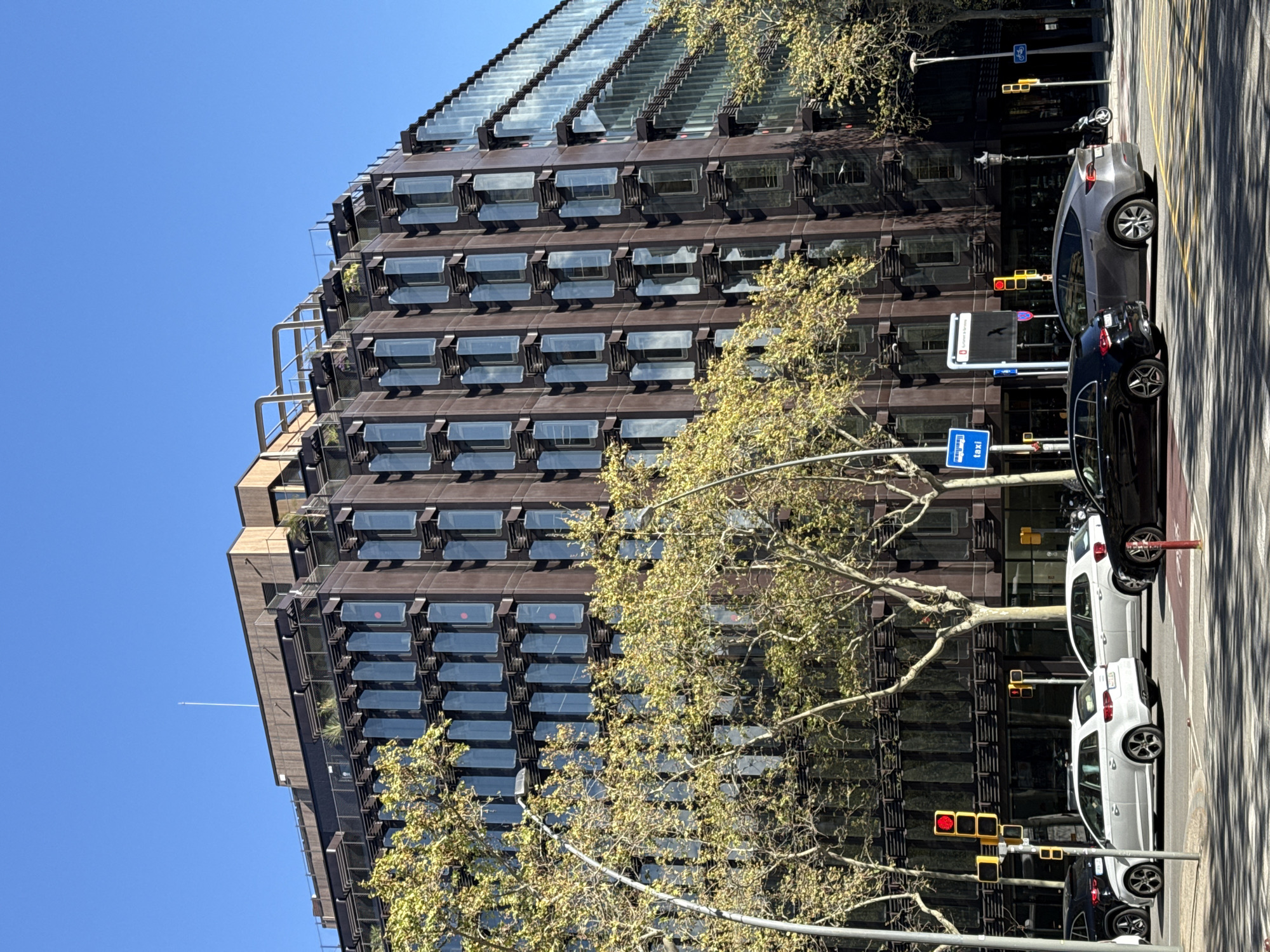 The image shows a portion of the Torre Llacuna office building in Barcelona, Spain. The building is characterized by its dark-colored, multi-story facade featuring rows of windows. A tree with budding green leaves partially obscures the building's lower levels. The street in front of the building features several parked cars, including a grey sedan, a black sedan, and a white compact car. Traffic lights are visible along the street, some displaying red signals. A taxi signpost is also present. The sky is clear and blue. The image is taken from a slightly low angle, looking upwards towards the building.