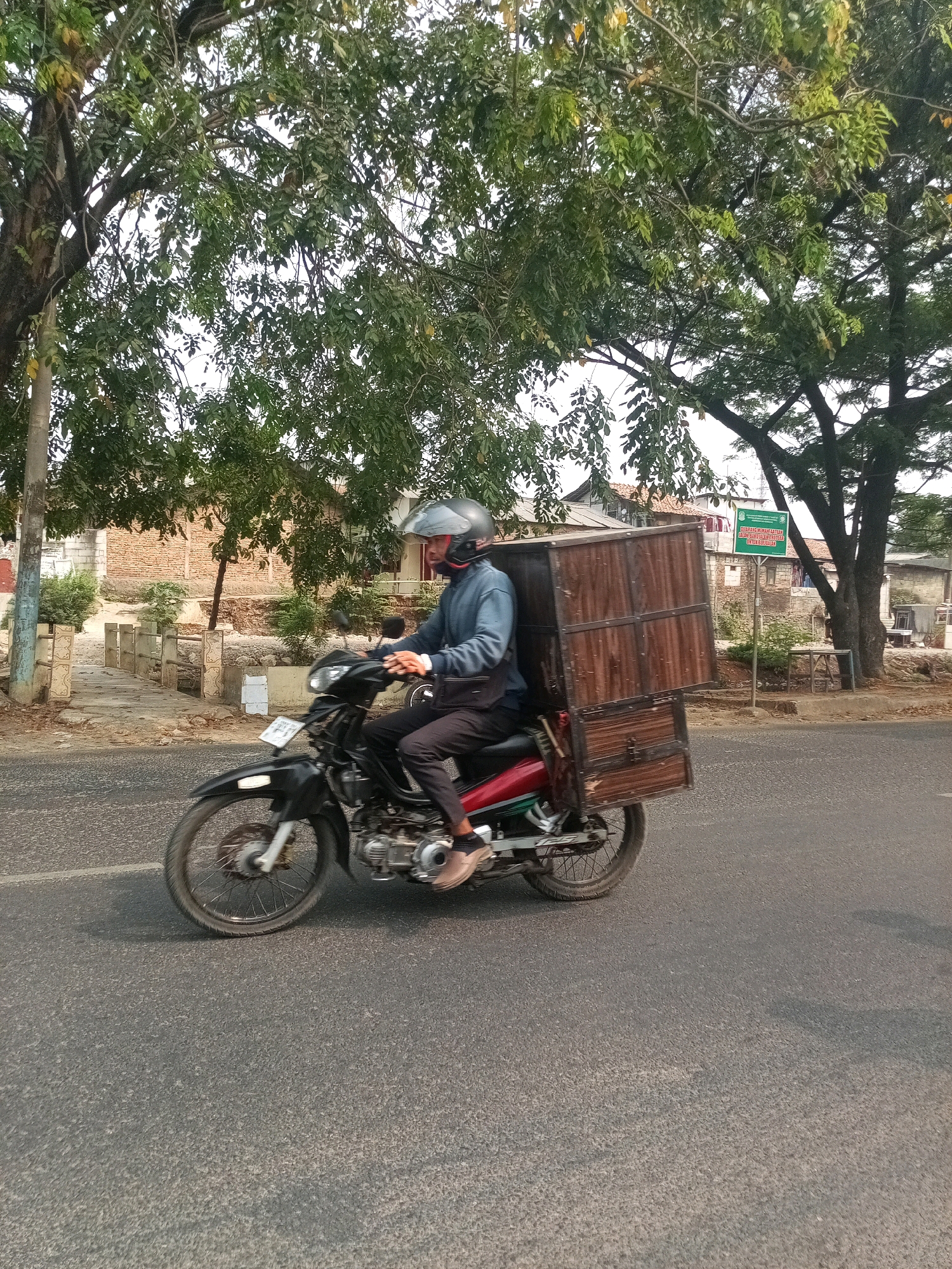 A man wearing a helmet rides a motorcycle with a large wooden box mounted on the rear. He is wearing a blue long-sleeved shirt and dark pants. The motorcycle is black with a red seat. The wooden box appears to be a delivery container. The man is riding on a paved road. In the background, there are trees, houses, and a green sign with Indonesian text. The scene appears to be in a residential or semi-rural area. The location is identified as Bekasi, Indonesia.