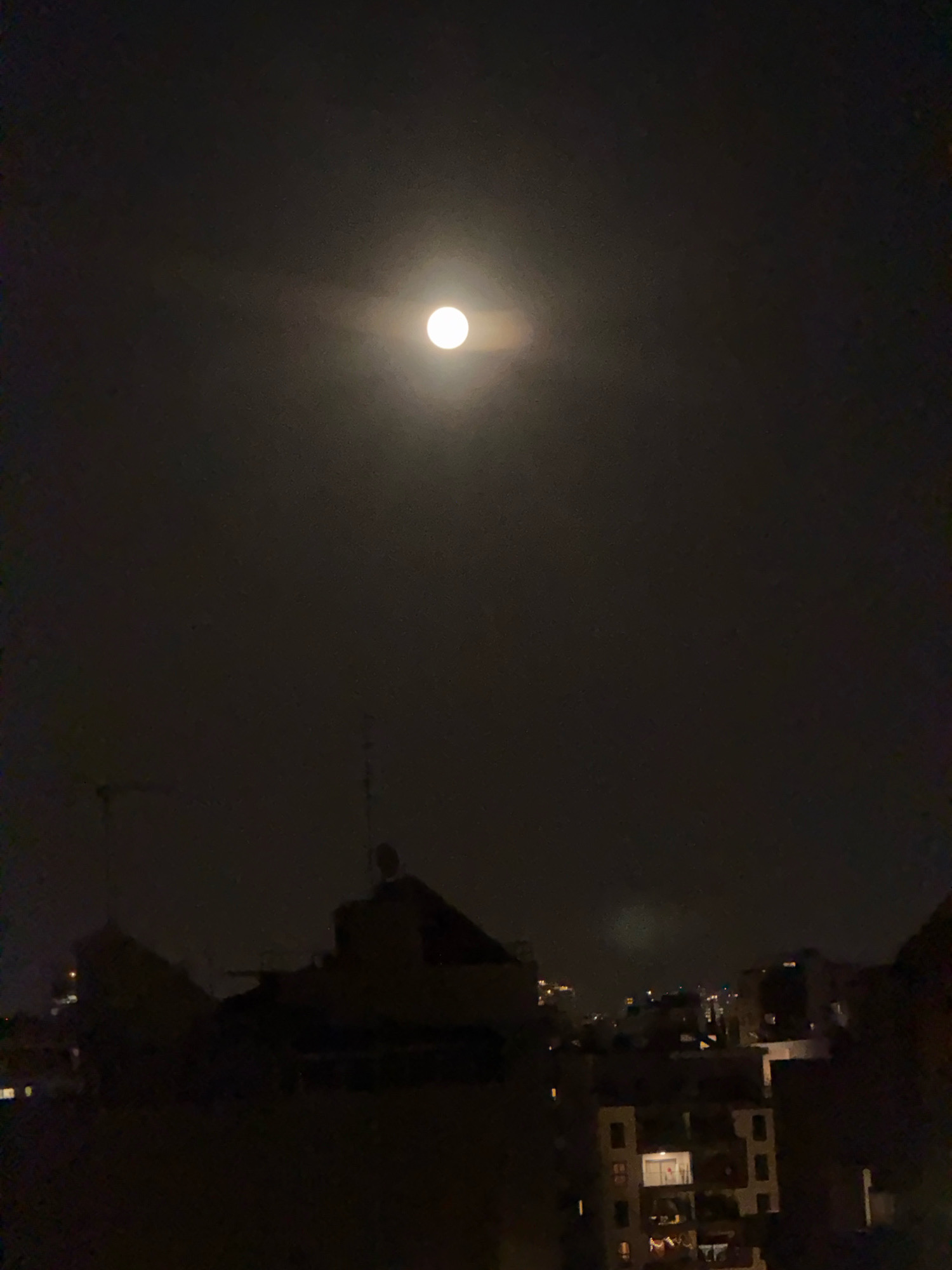The image shows a night view of a city skyline, likely Ramat Gan, Israel, under a full moon. The moon is prominently positioned in the upper center of the frame, appearing as a bright white orb surrounded by a soft halo of light, indicating it is partially obscured by thin clouds. Below the moon, the silhouette of city buildings is visible. The foreground is dominated by the dark shapes of rooftops and building structures, with some windows lit from within, suggesting indoor activity. In the mid-ground and background, more buildings are discernible, forming a dense urban landscape. The overall atmosphere is dark and nocturnal, with the moon being the primary light source in the sky. The image appears to be taken from an elevated vantage point, looking out over the city.