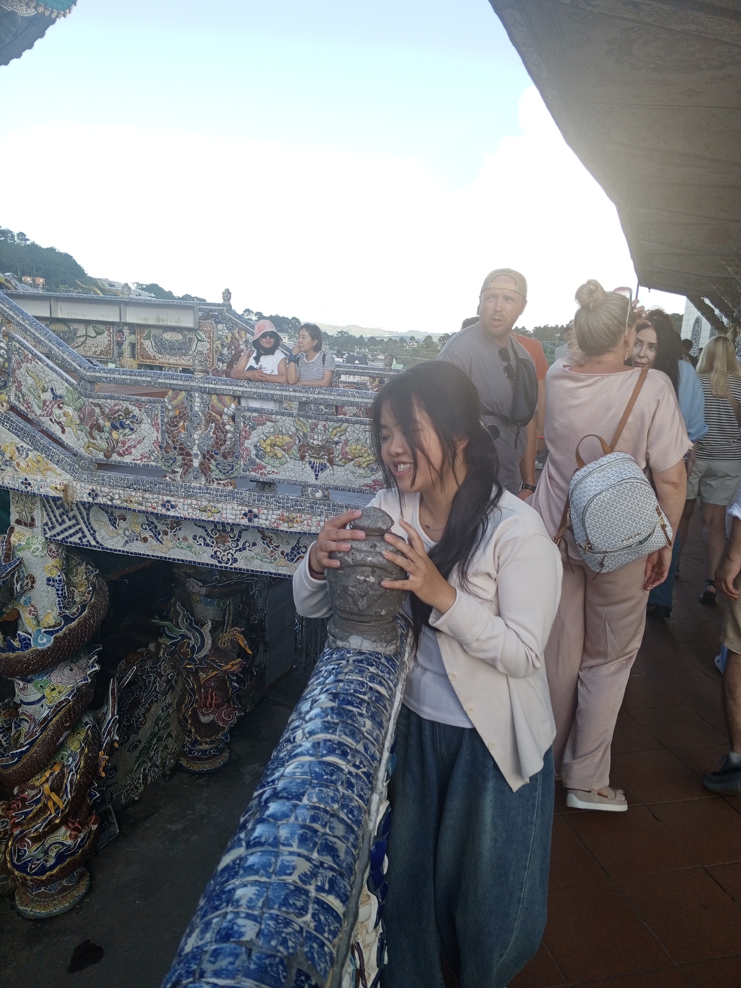 In Xuân Thành, Vietnam, a woman with long dark hair and a light-colored cardigan stands in the foreground, leaning against a ornate blue and white tiled railing. She is looking down and to her left, her hands resting on the railing. Behind her, a woman in a pale pink outfit with a small patterned backpack walks away from the camera, while other individuals are visible in the background. The scene appears to be an outdoor tourist area, possibly a temple or historical site, characterized by intricate mosaic decorations on a structure to the left of the frame, featuring mythological creatures like dragons. The sky is bright and overcast, suggesting daytime.