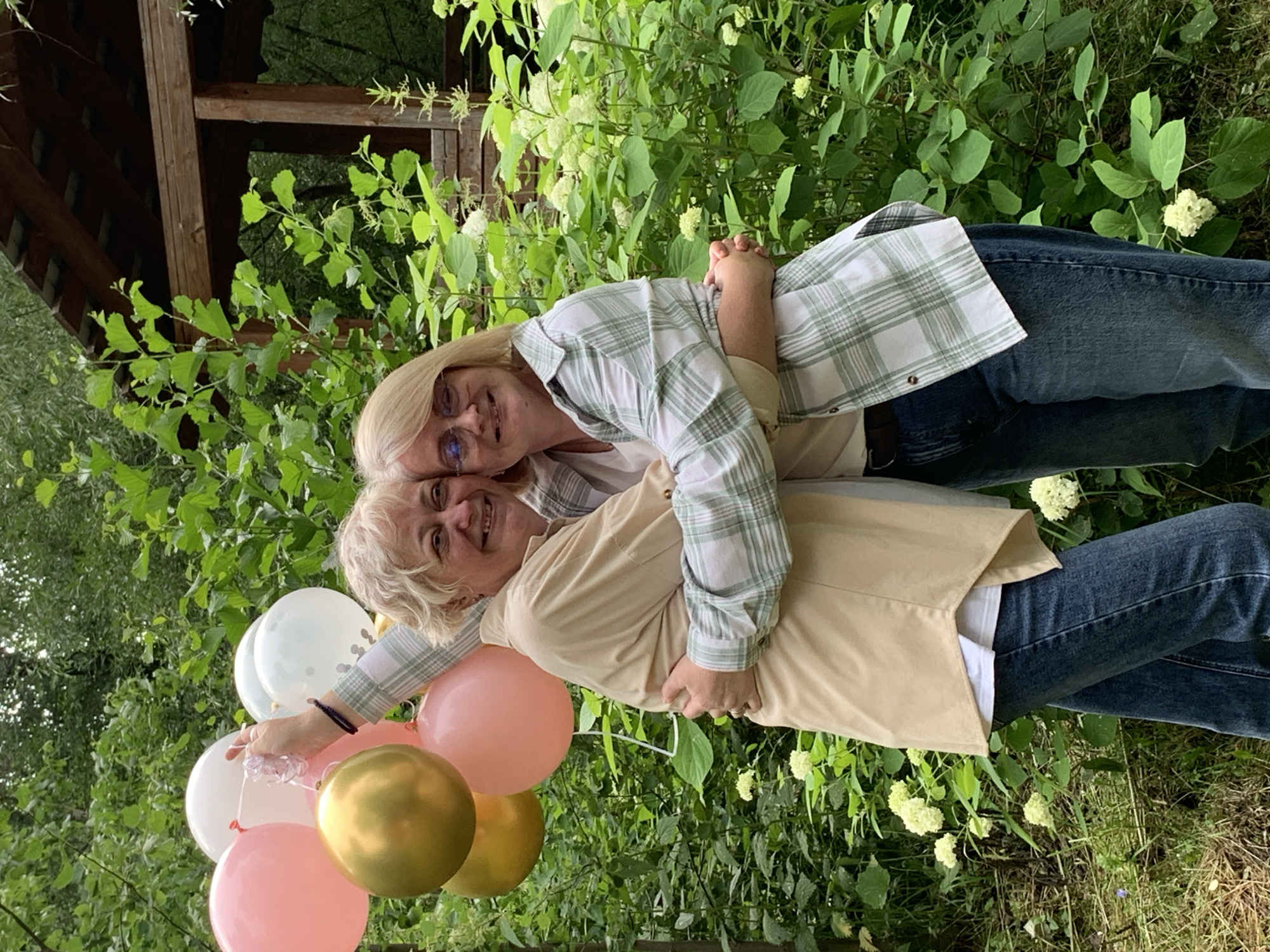 This media file shows two women embracing outdoors, with a backdrop of lush green foliage and a wooden structure. The woman on top is wearing a green and white plaid shirt and blue jeans, with blonde hair and glasses. She is smiling and holding onto the woman below her. The woman below has blonde, curly hair and is wearing a tan jacket over a white shirt and blue jeans. She is also smiling.
Several balloons are visible in the foreground, including pink, gold, and white ones. The scene suggests a celebratory or special occasion, perhaps a birthday or anniversary, given the balloons and the affectionate embrace. The lighting indicates it is daytime, with soft natural light illuminating the subjects and their surroundings. The greenery suggests a garden or park setting. The overall mood conveyed is joyful and warm. No visible text is present in the image. The location context is Chaikovsky, Russia.