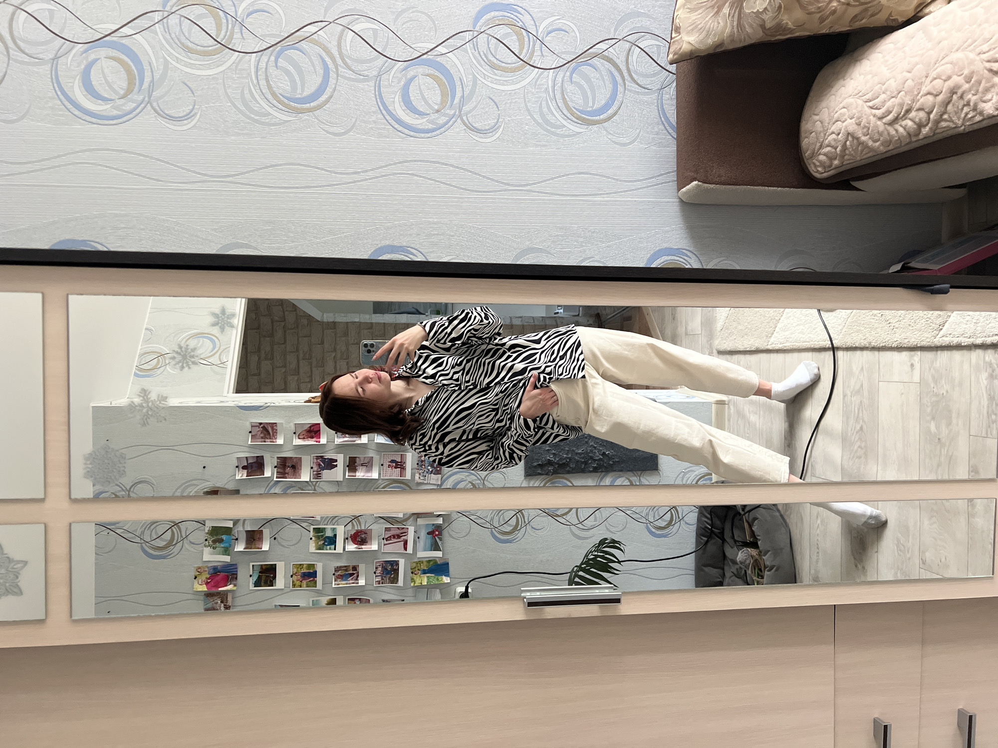 A woman is captured in a full-body mirror selfie, standing upright indoors. She has dark hair and is wearing a black and white zebra-print long-sleeved shirt paired with light-colored, possibly beige, trousers and white socks. She holds a smartphone to her ear or is looking at its screen, with a neutral expression.
The mirror appears to be composed of multiple rectangular panels, possibly part of a wardrobe or a decorative wall. Reflected in the mirror and visible above it is wallpaper featuring a light background with abstract circular and wavy patterns in blue, grey, and tan tones. On some of the mirror panels, small instant photos, likely personal snapshots, are affixed in rows. Decorative elements resembling snowflakes or floral designs are also visible on some mirror sections.
The floor, visible at the bottom of the image and in reflection, is light-colored wood laminate, partially covered by a light-toned rug. To the woman's right in the reflection, a segment of a brown upholstered sofa or bed with light cushions is visible. A black electrical cord runs across the floor. Other reflections include what might be a brick-patterned wall and a small potted plant. The setting suggests a domestic environment, possibly a bedroom or living room in Chaikovsky, Russia. There is no visible text in the image.