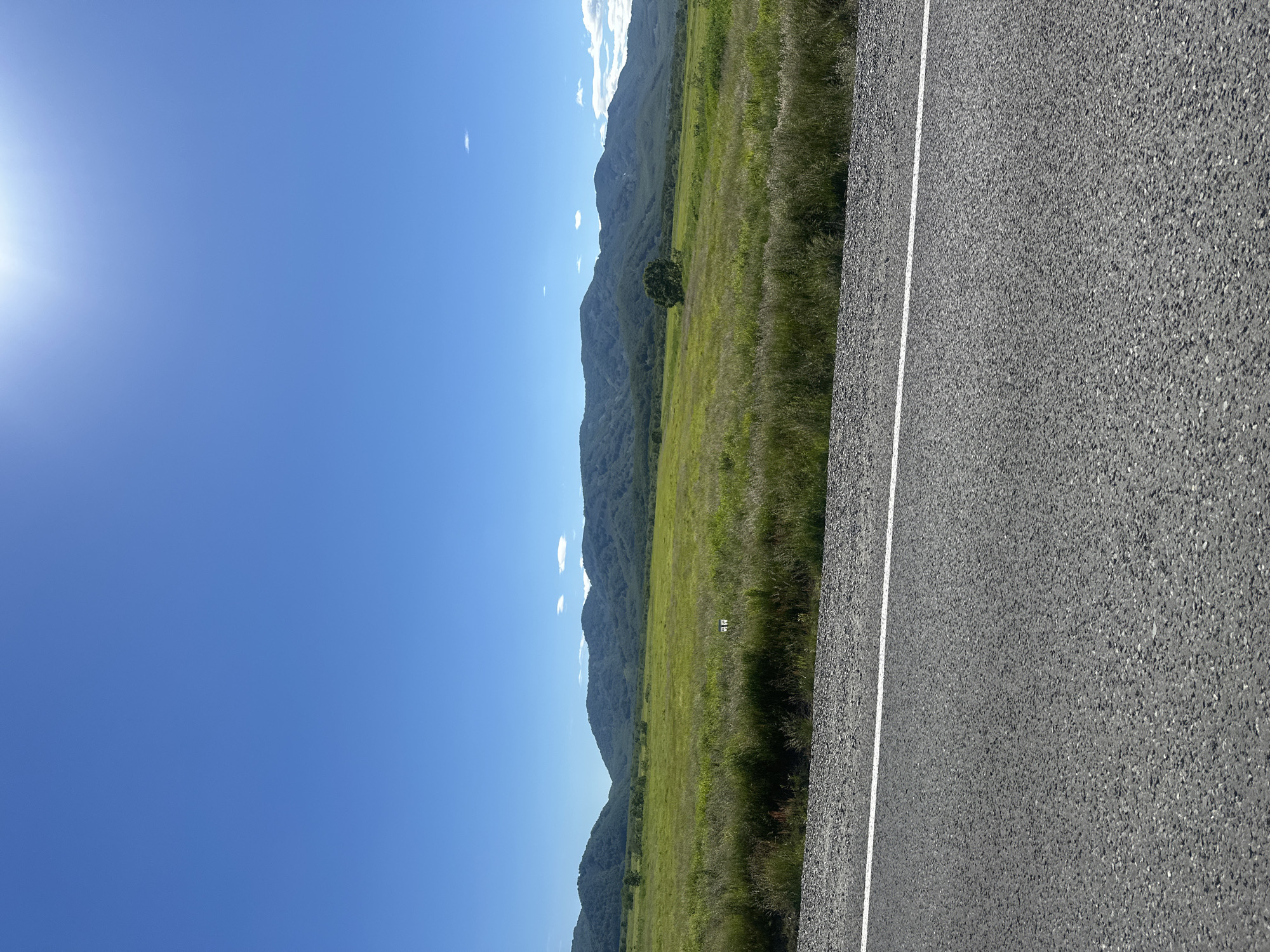The image depicts a wide, scenic view of a grassy plain with rolling, tree-covered mountains in the background under a clear blue sky with some scattered white clouds. In the foreground, a paved road with a white line marking its edge runs horizontally across the bottom of the frame. The road appears to be asphalt with visible gravel. To the right of the road and slightly into the grassy field are two small, white posts with some dark markings on them, possibly indicating a boundary or a sign. A lone tree stands in the middle of the grassy field. The overall impression is a rural landscape. The prompt requests to identify the location as Petropavlovsk-Kamchatsky, Russia; however, there are no visual cues within the image itself that can confirm this specific location.